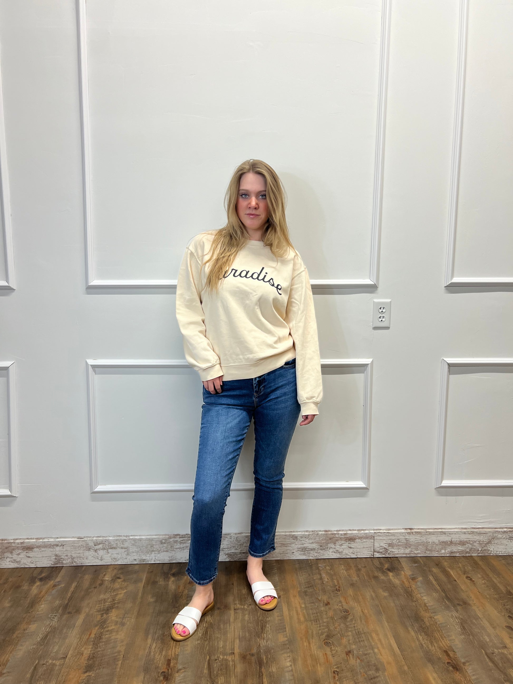 Person wearing a beige sweatshirt and blue jeans standing in front of a white door.