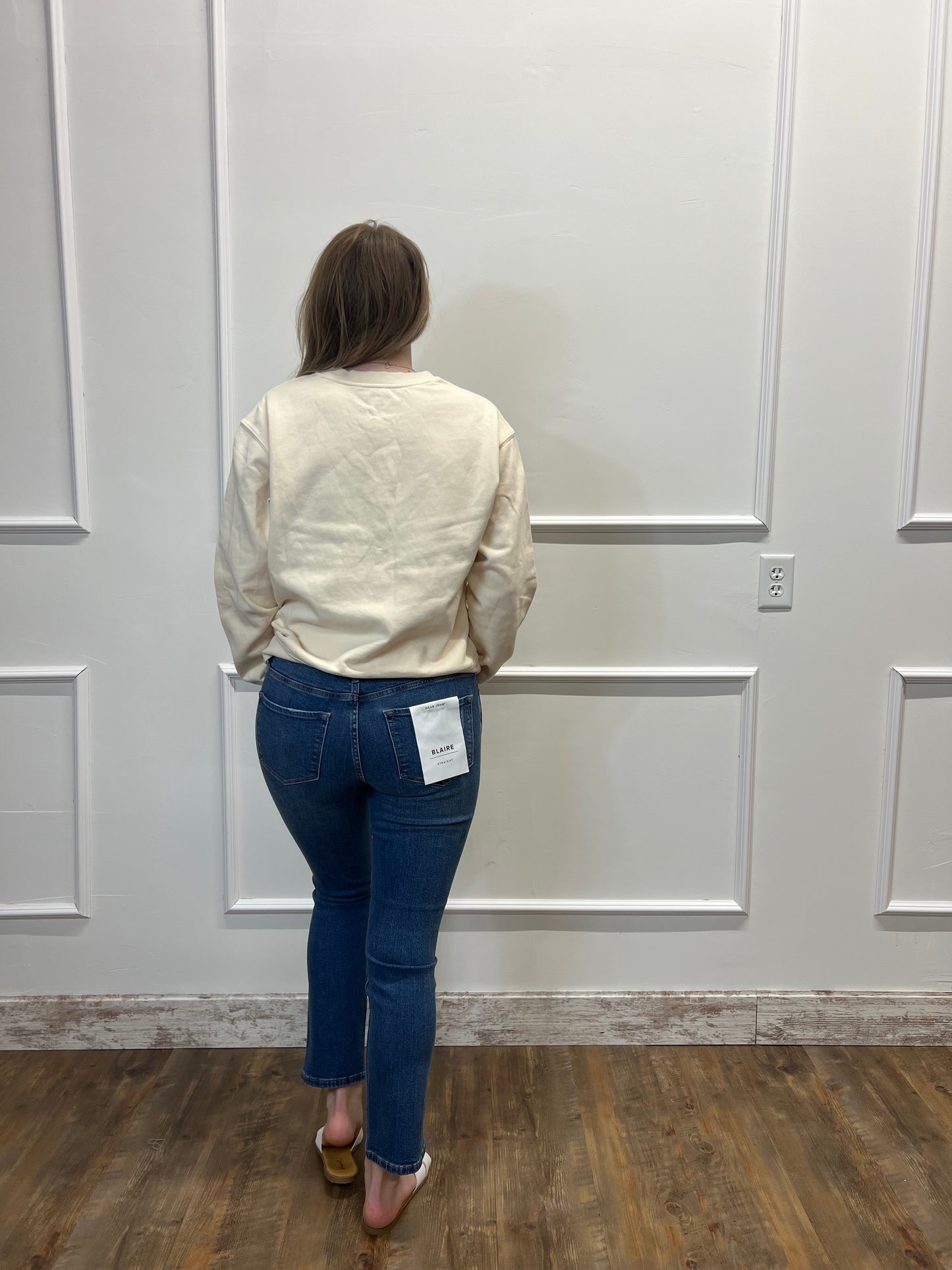 Person wearing a beige jacket and blue jeans standing in front of a white door.