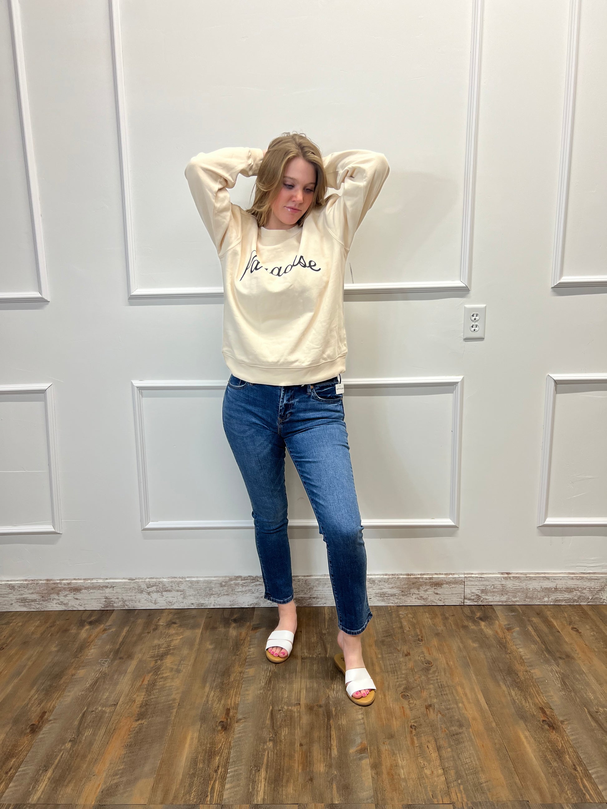 Person wearing a beige hoodie and blue jeans standing in a room with wooden flooring and white walls.