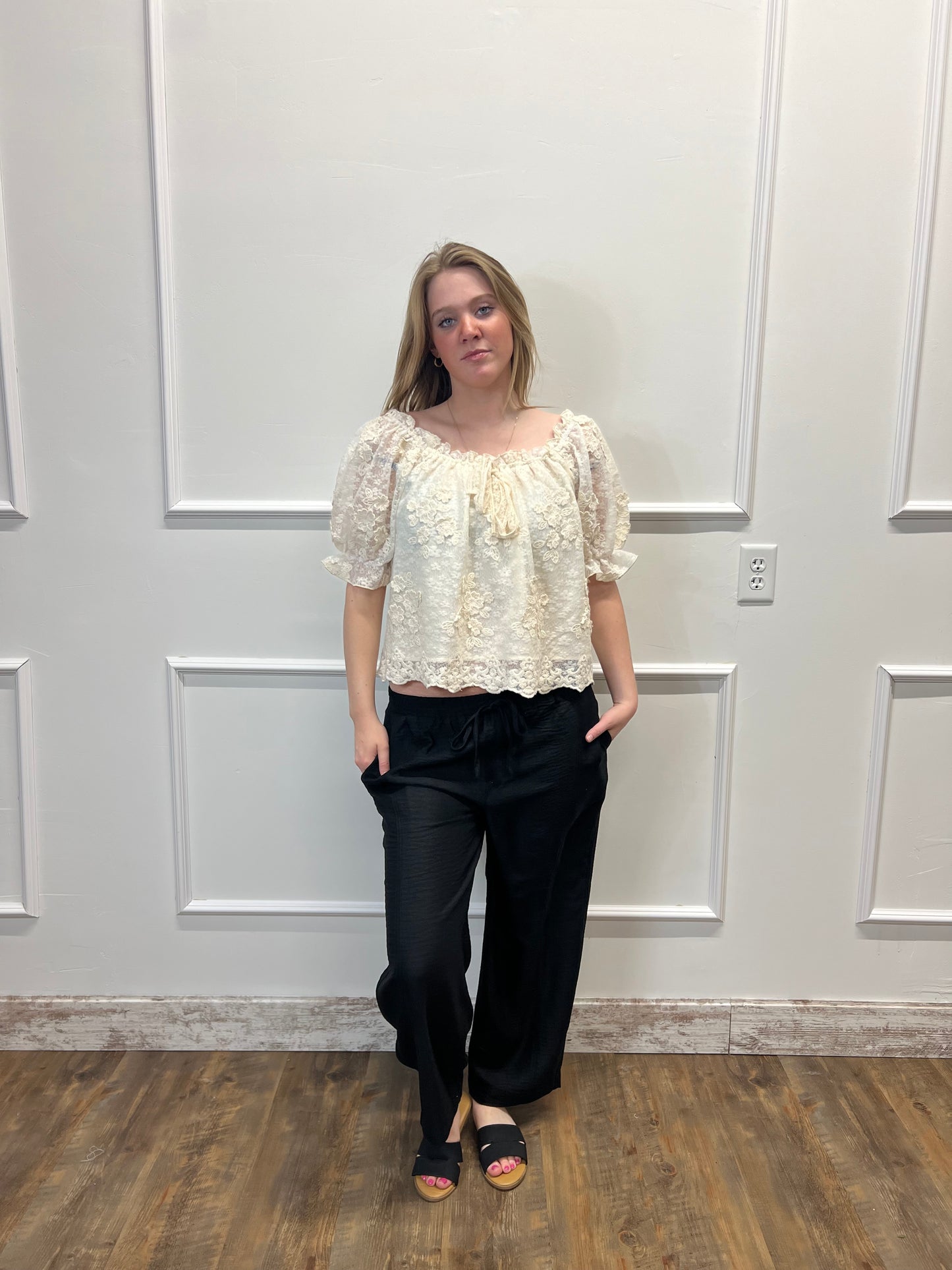 Person wearing a white lace top and black pants standing in front of a white paneled wall.