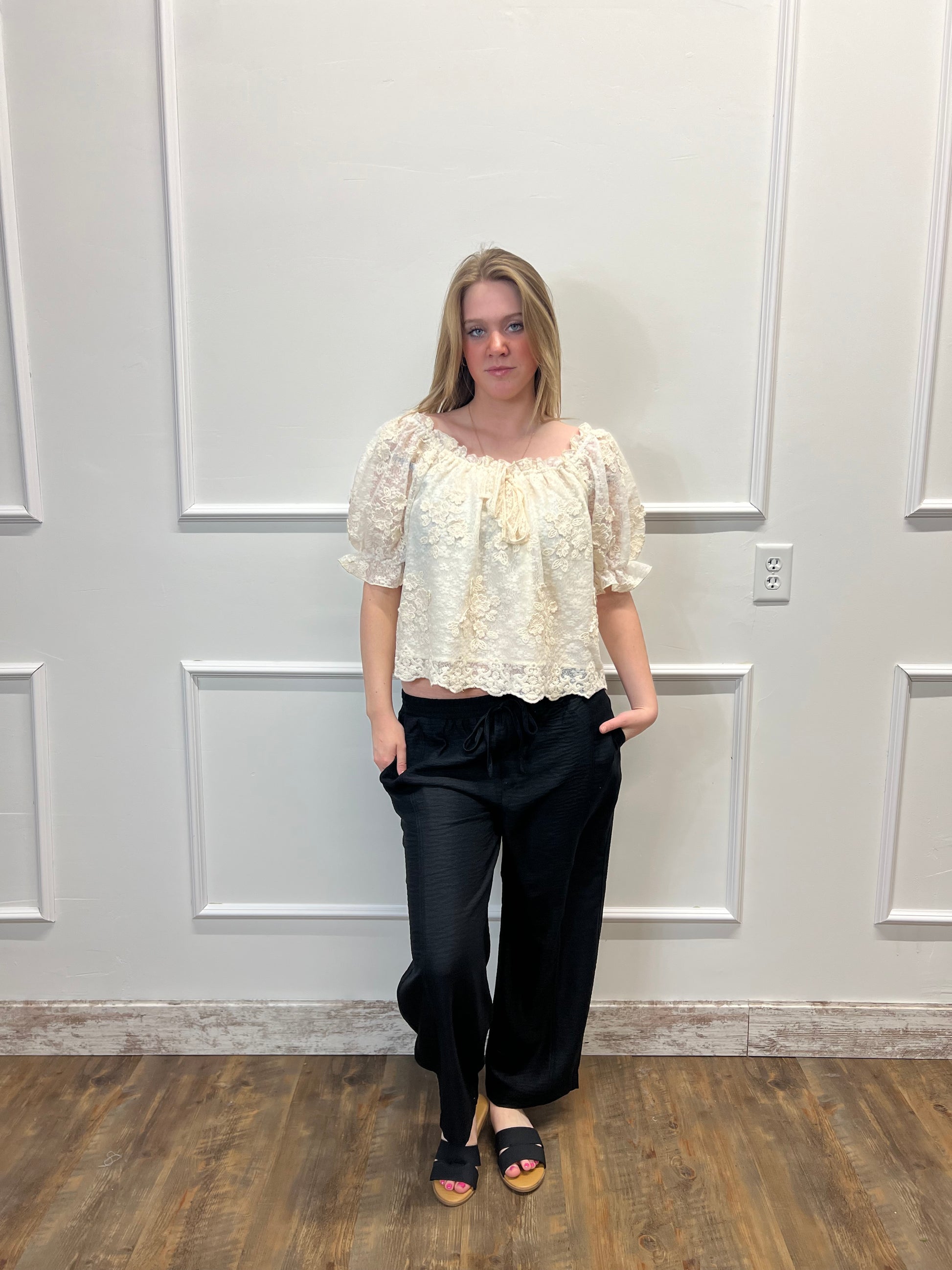 Person wearing a white lace top and black pants standing in front of a white door.