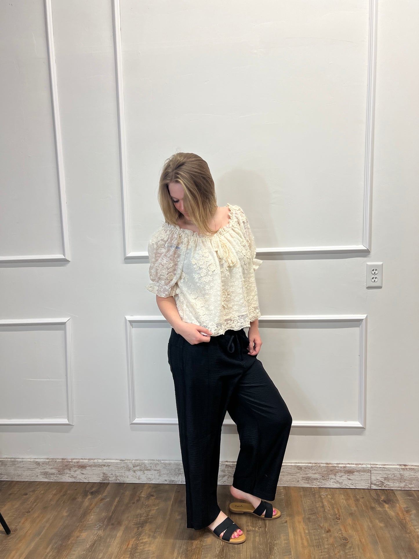 Person wearing a white lace top and black pants standing in front of a white paneled wall.