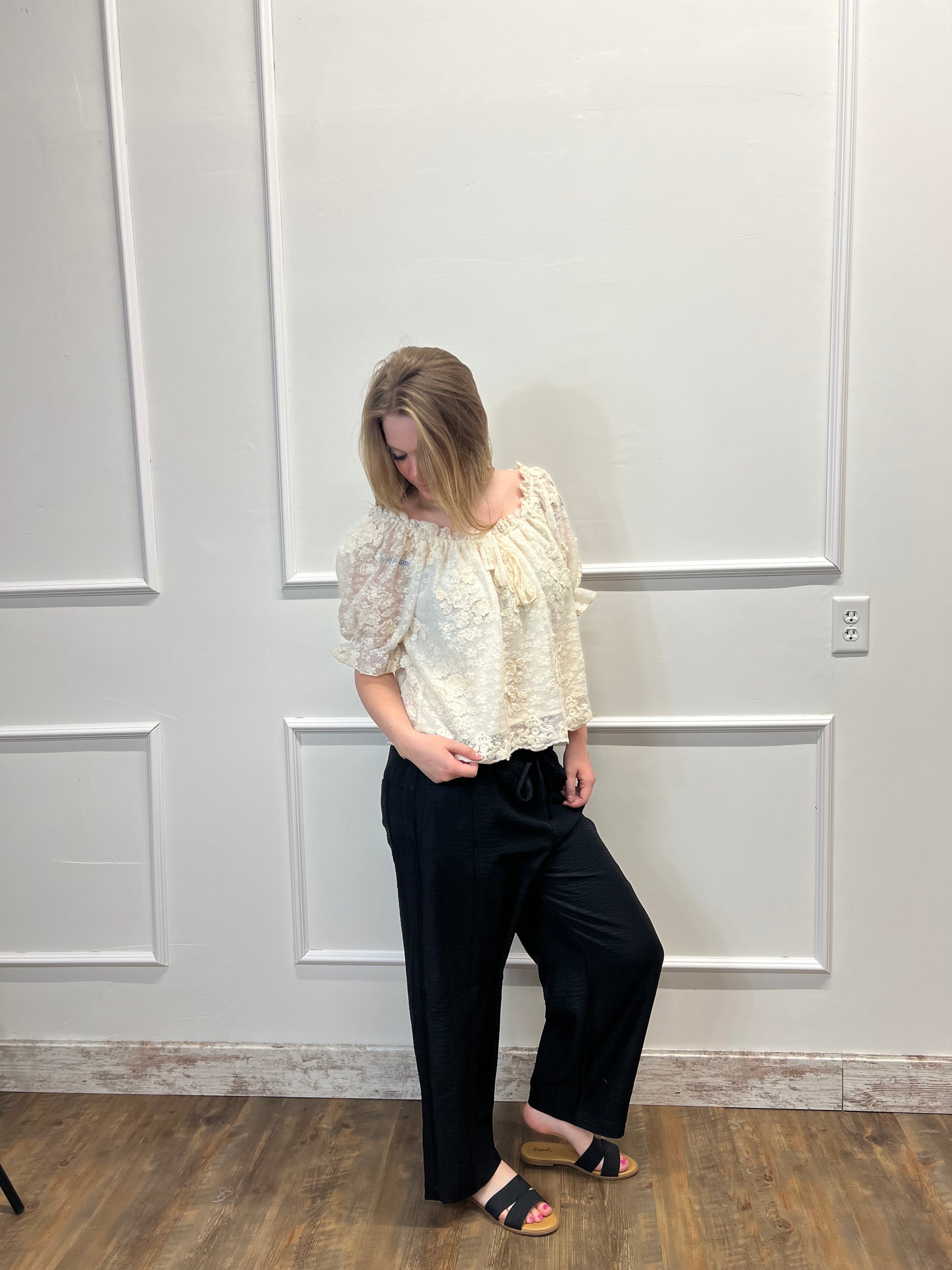 Person wearing a white lace top and black pants standing in front of a white paneled wall.