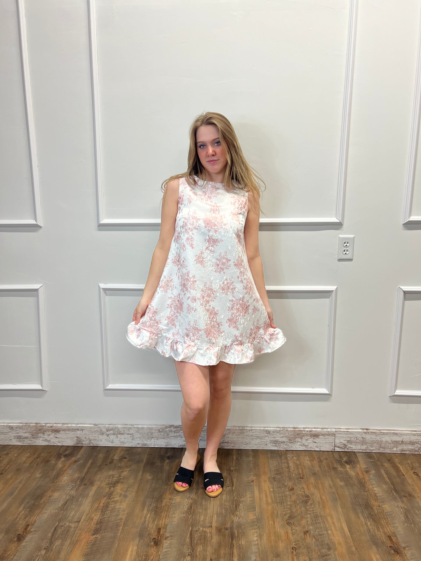 Woman wearing a floral dress standing in a room with wooden flooring and white walls.