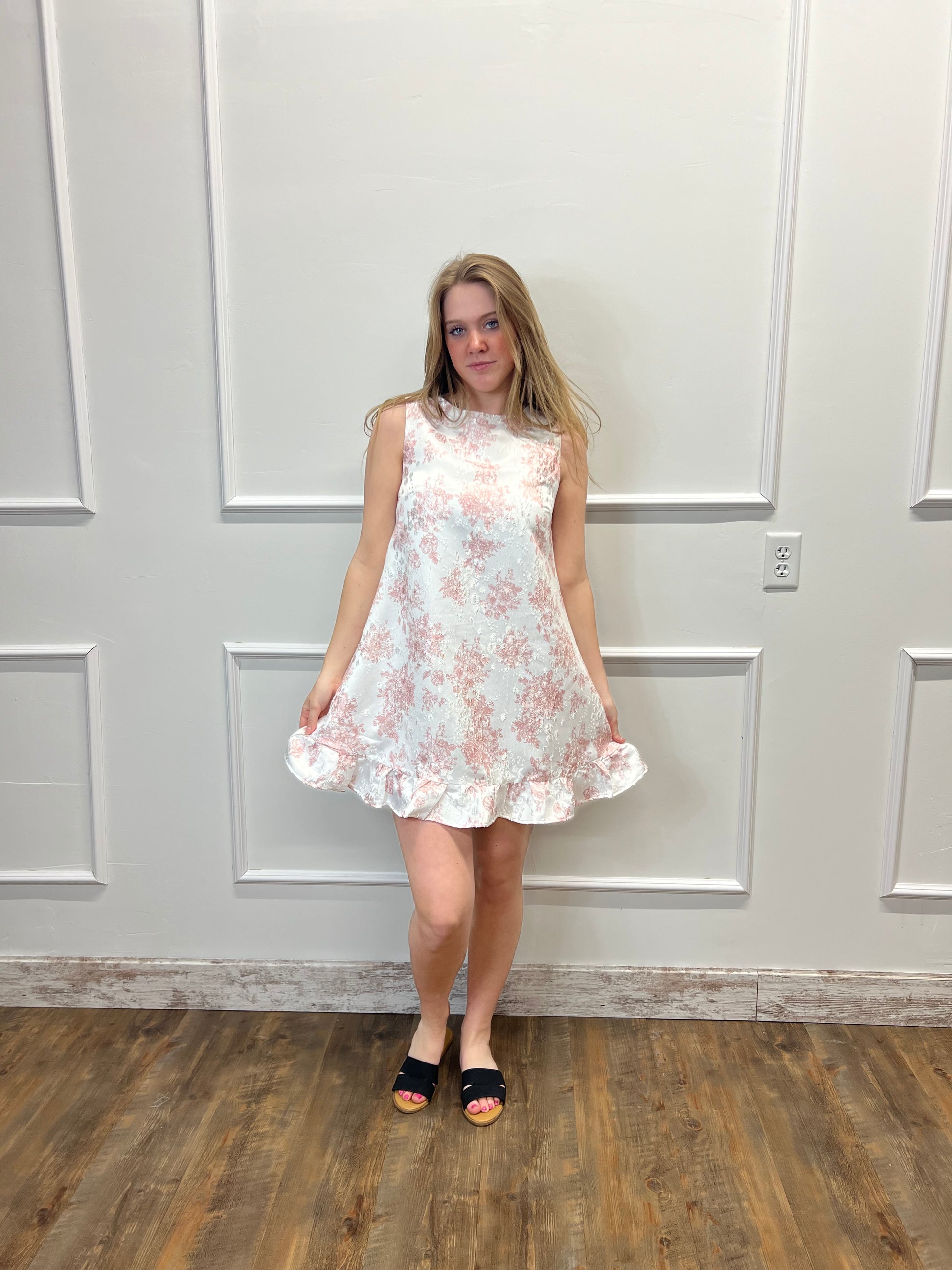 Woman wearing a floral dress standing in a room with wooden flooring and white walls.