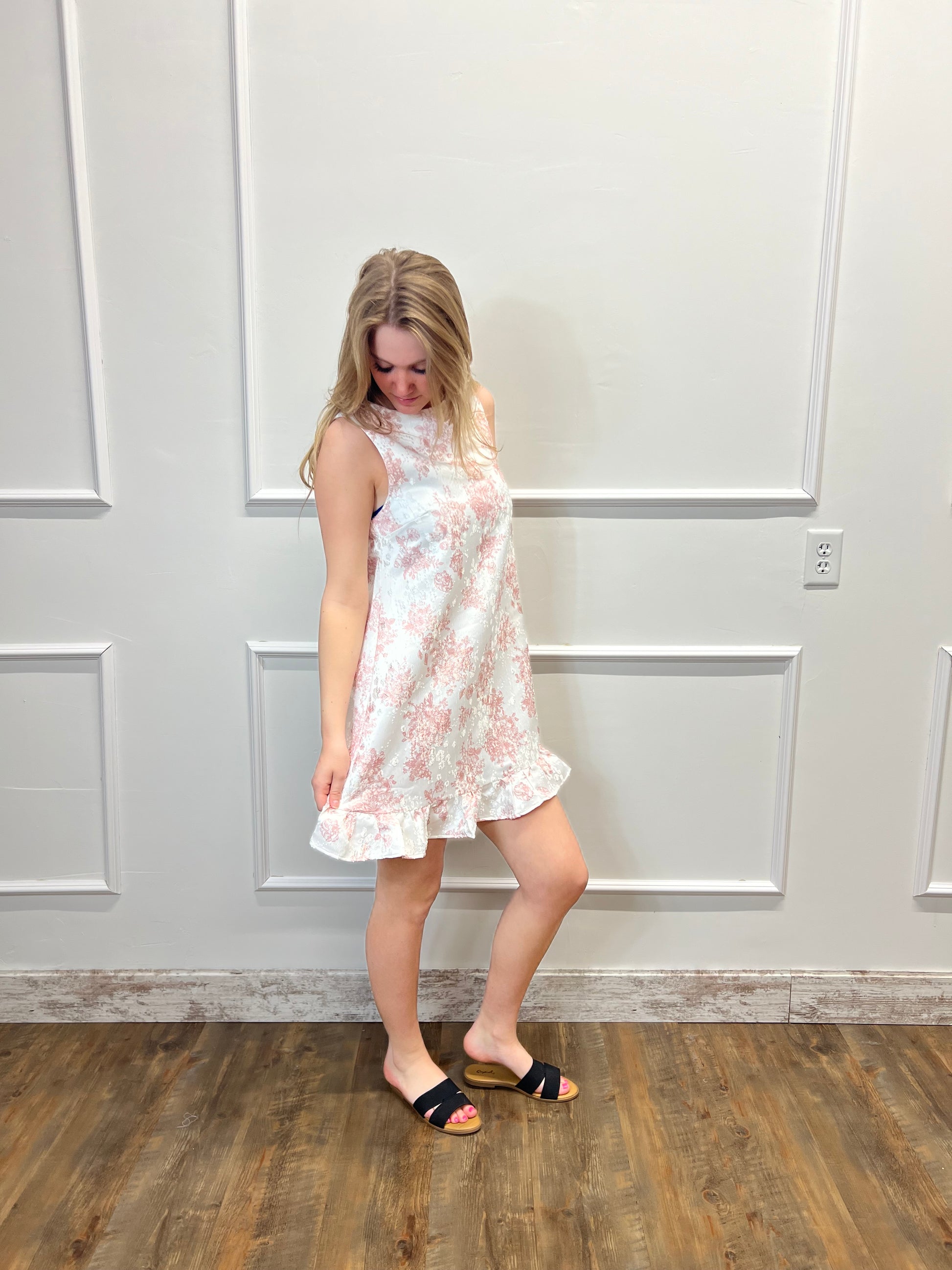 Woman in a floral dress standing against a white paneled wall.