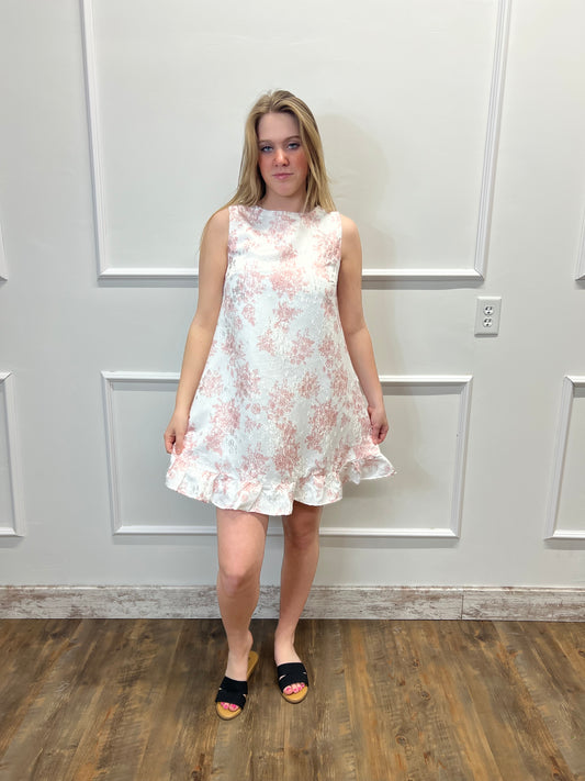 Woman wearing a floral dress standing in a room with wooden flooring and white walls.