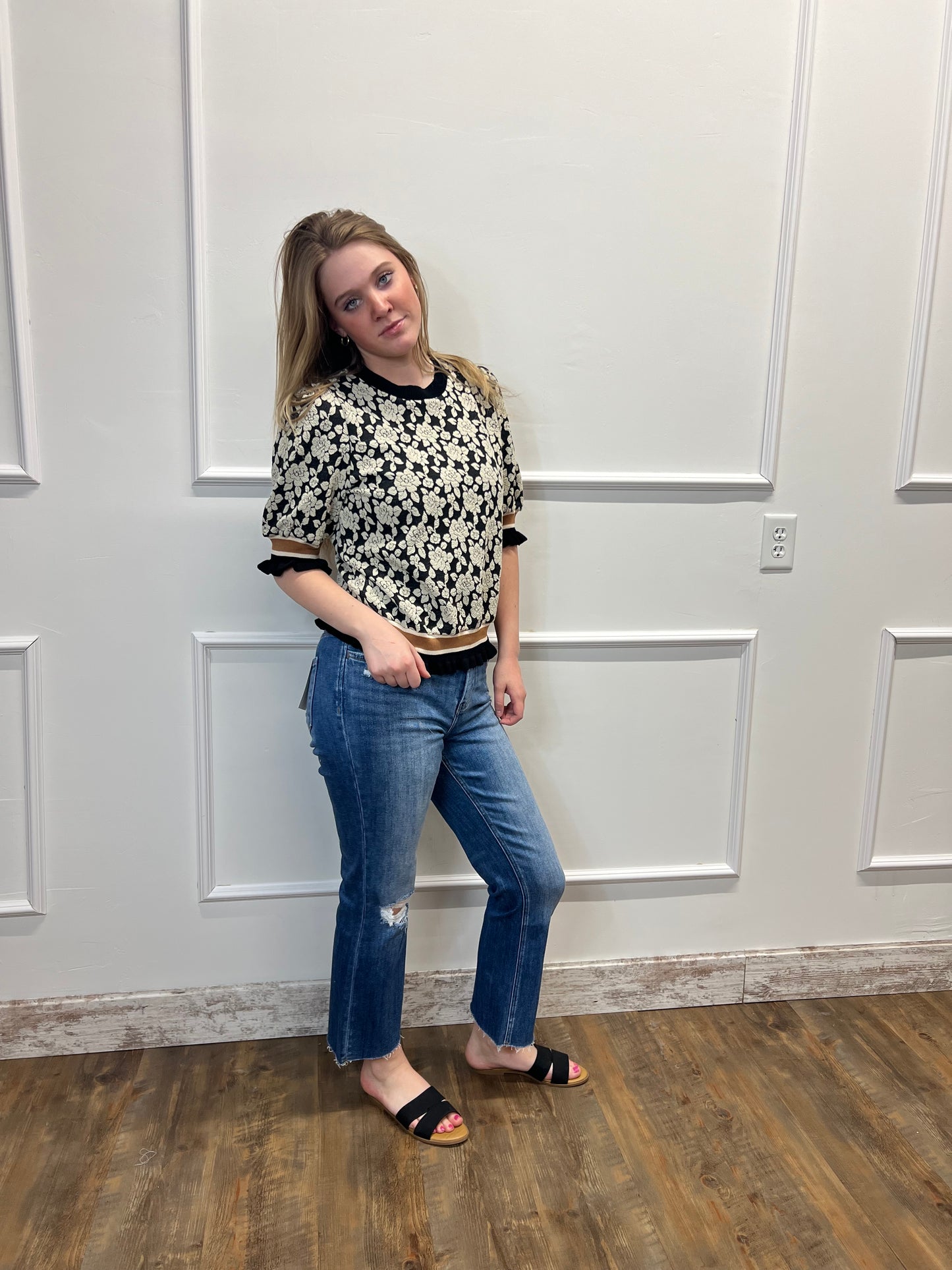 Person wearing a patterned top and jeans standing in front of a white door.