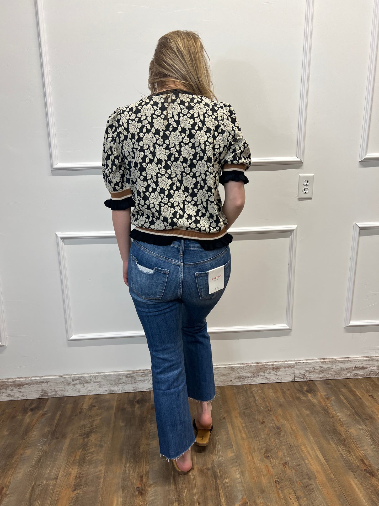 Person wearing a floral-patterned jacket and blue jeans standing in front of a white door.