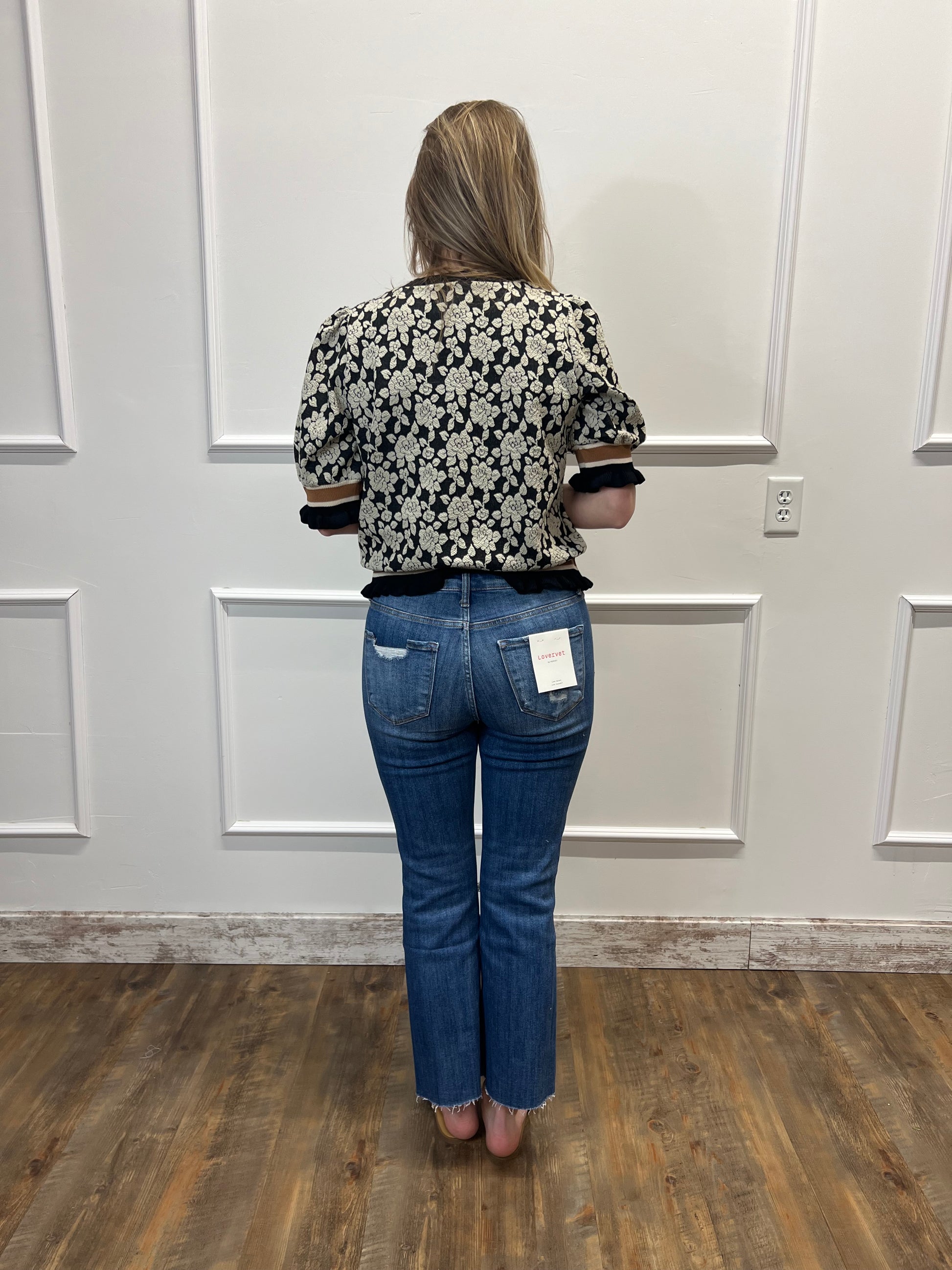Person wearing a floral shirt and blue jeans standing in front of a white door.