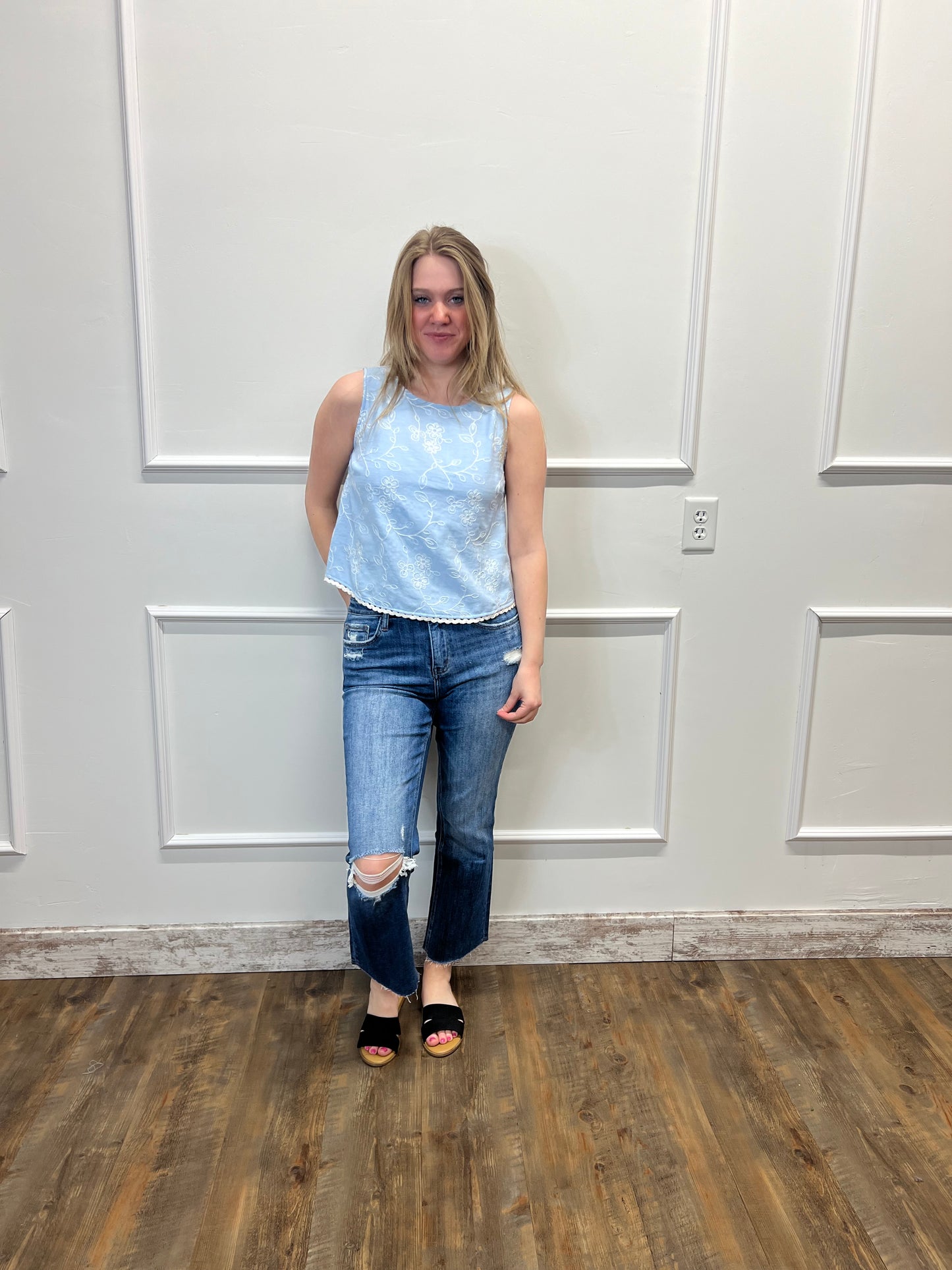 Person wearing a light blue sleeveless top and ripped jeans standing in a room with a white paneled wall and wooden floor.