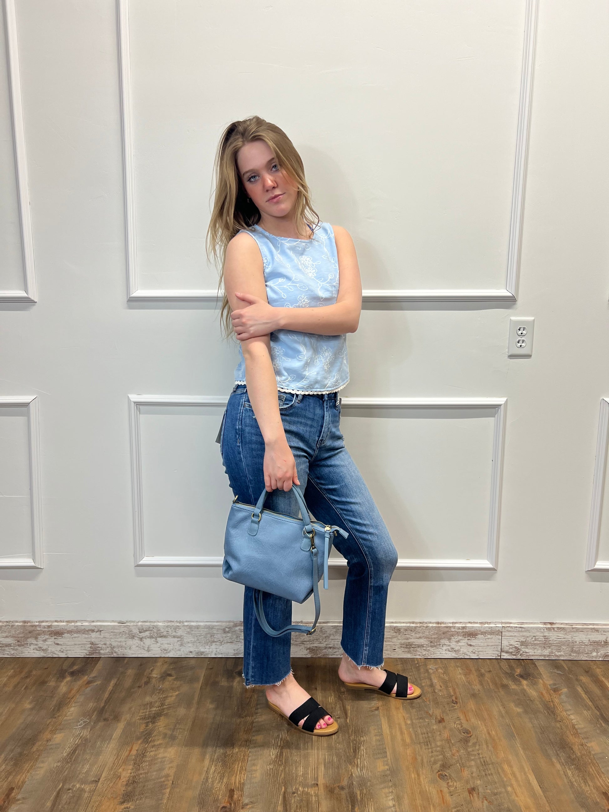 Person wearing a light blue sleeveless top and jeans, holding a blue handbag, standing against a white wall.