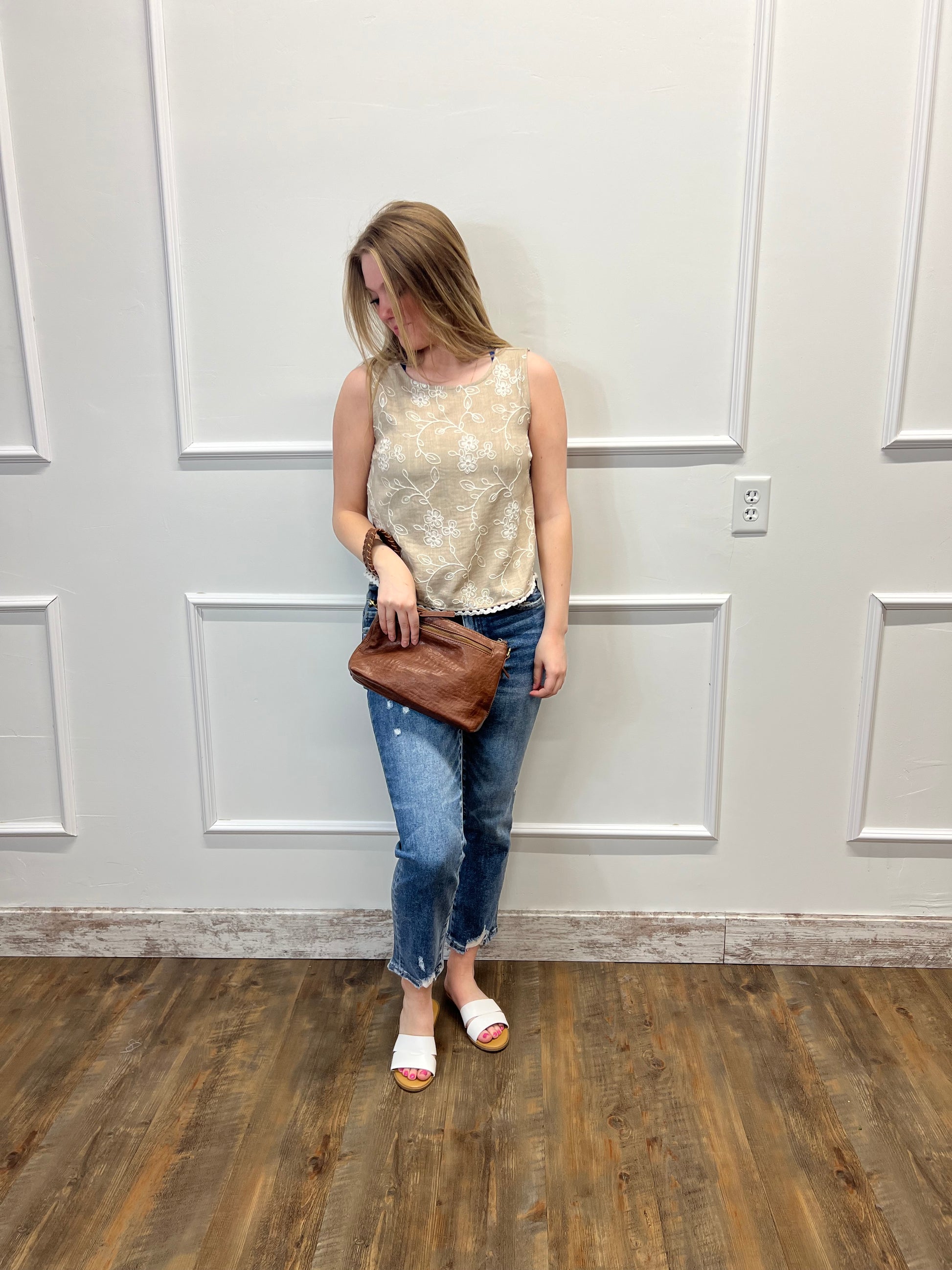 Person wearing a patterned top, brown jacket, and blue jeans standing against a white paneled wall.