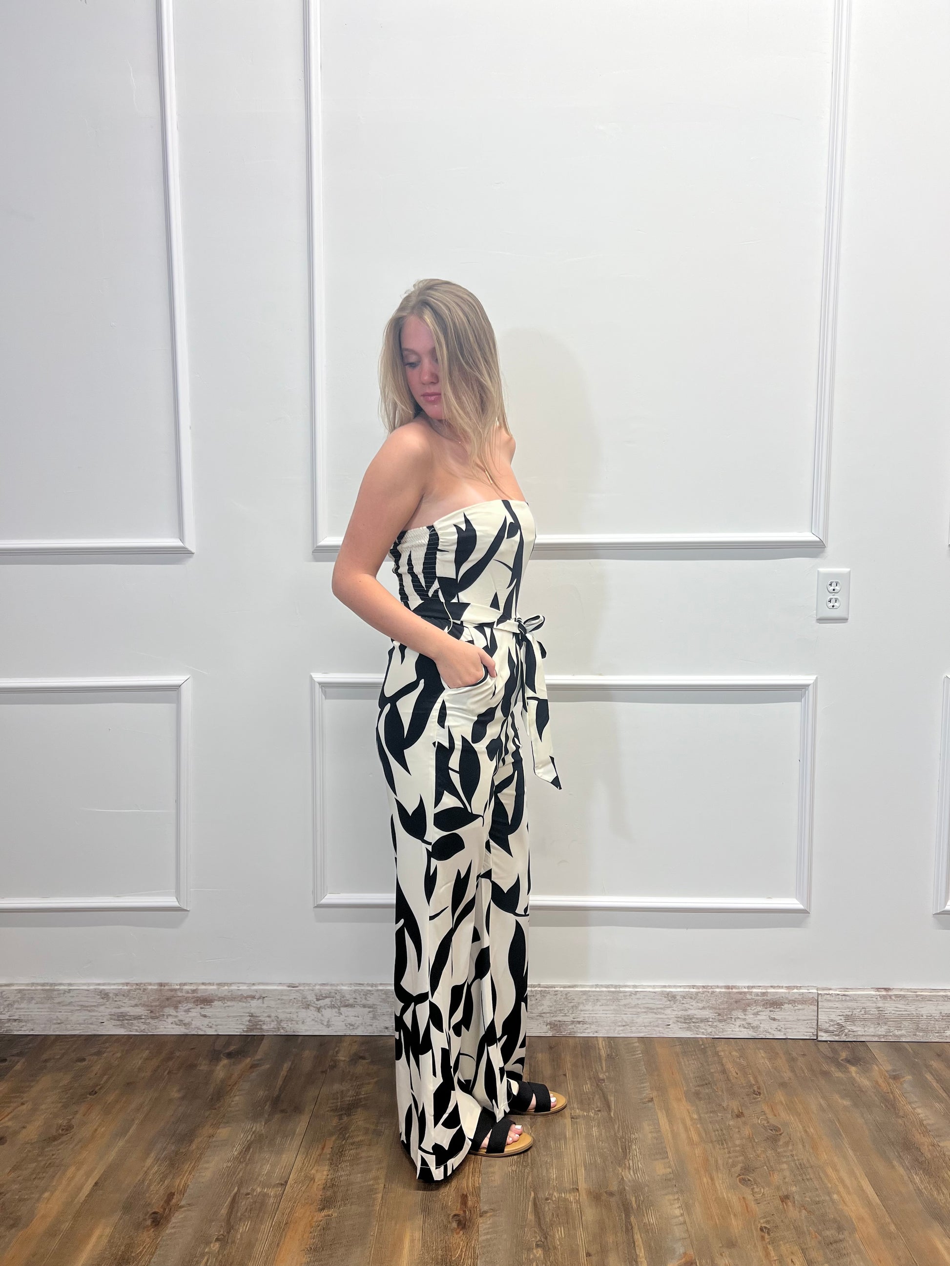 Woman wearing a strapless black and white patterned jumpsuit against a white paneled wall.