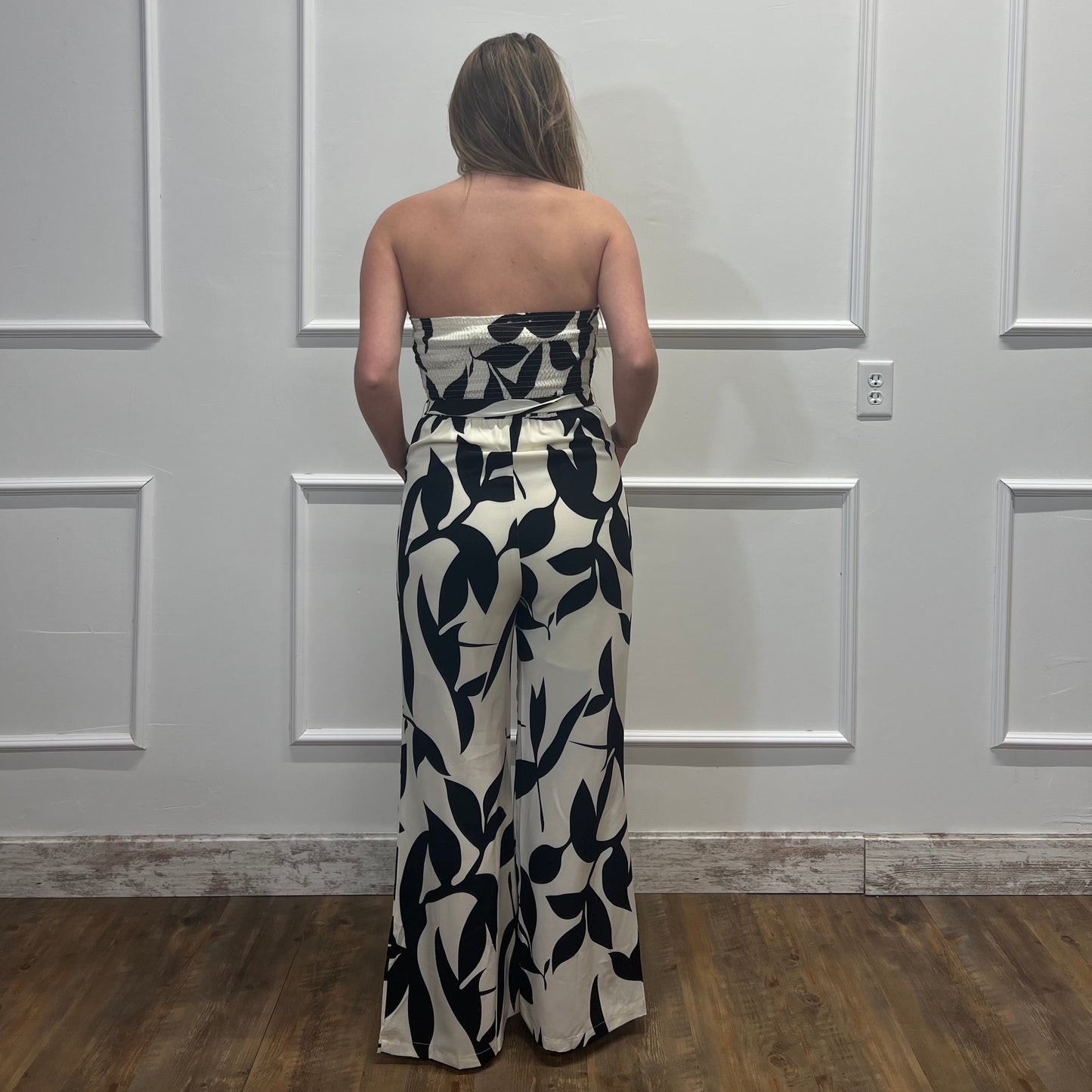 Person wearing a strapless black and white floral jumpsuit standing against a white paneled wall.