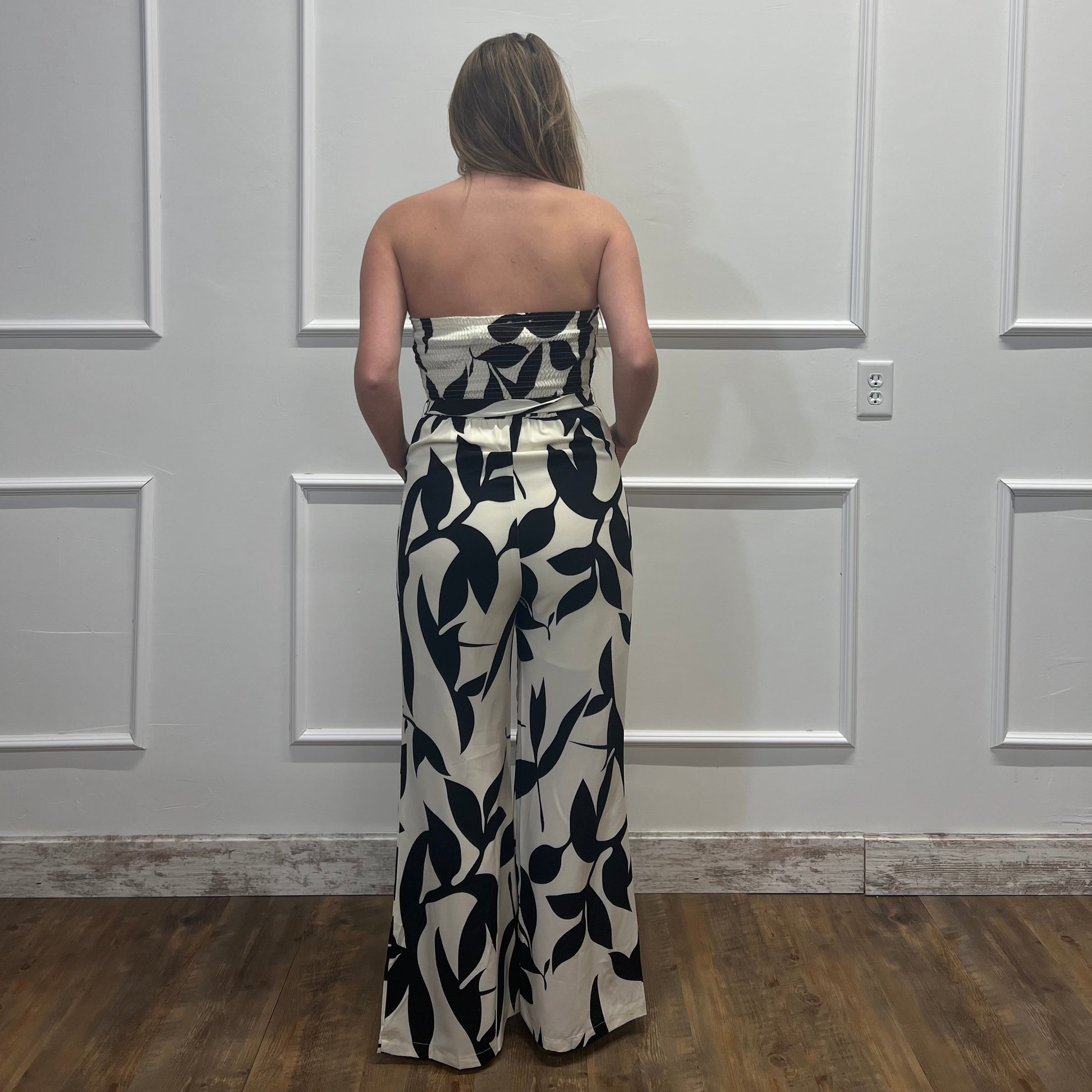 Person wearing a strapless black and white floral jumpsuit standing against a white paneled wall.