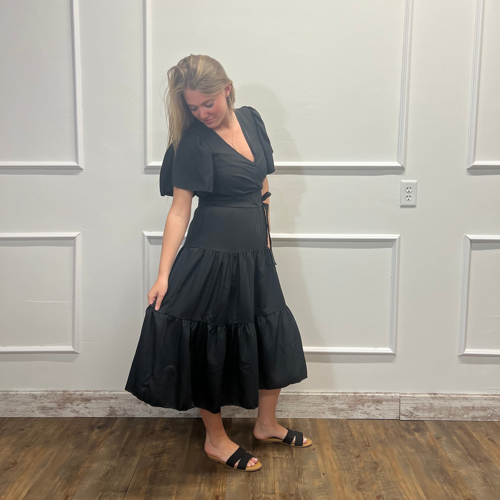 Person wearing a black dress standing in front of a white paneled wall.