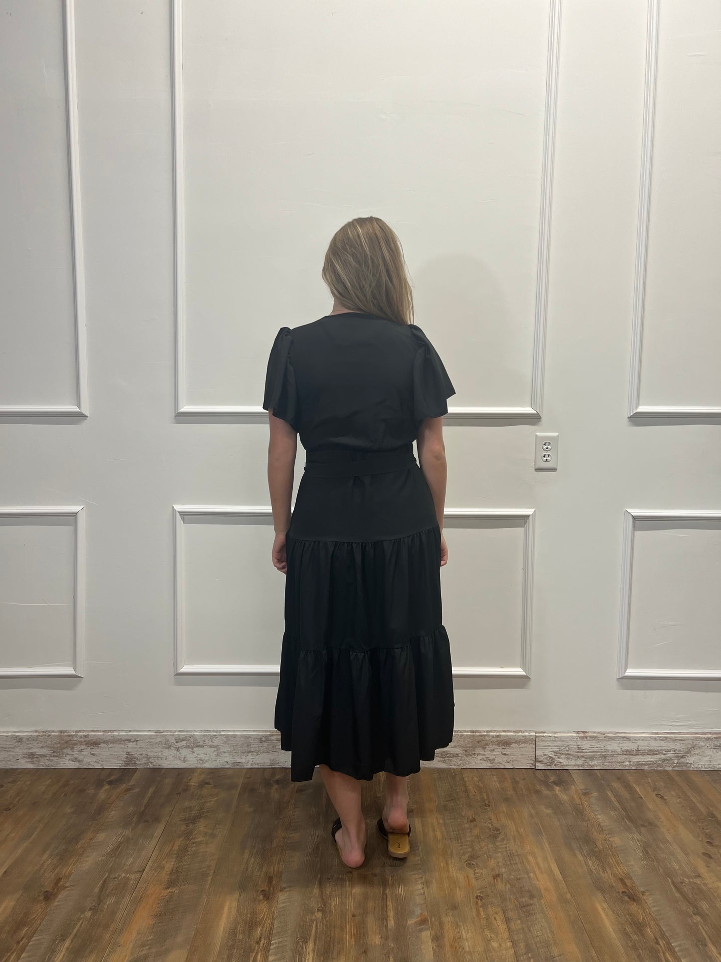 Person wearing a black dress standing against a white paneled wall.