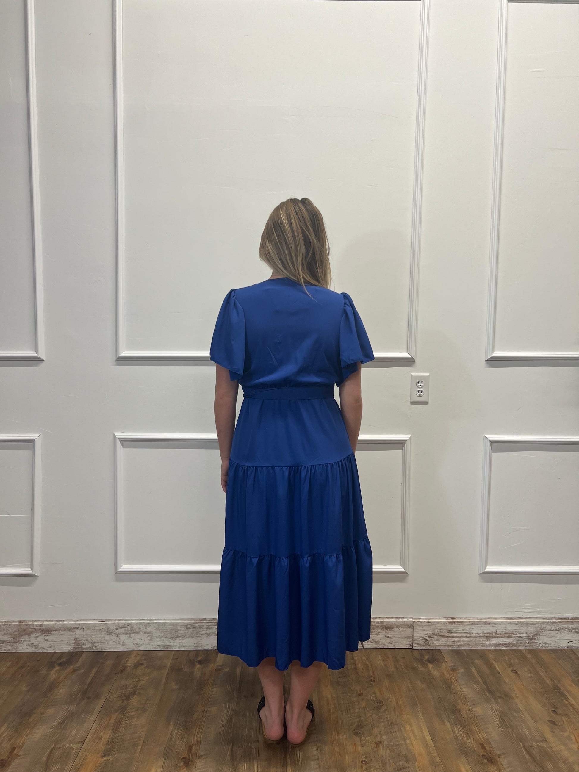 Woman wearing a blue dress standing against a white paneled wall.