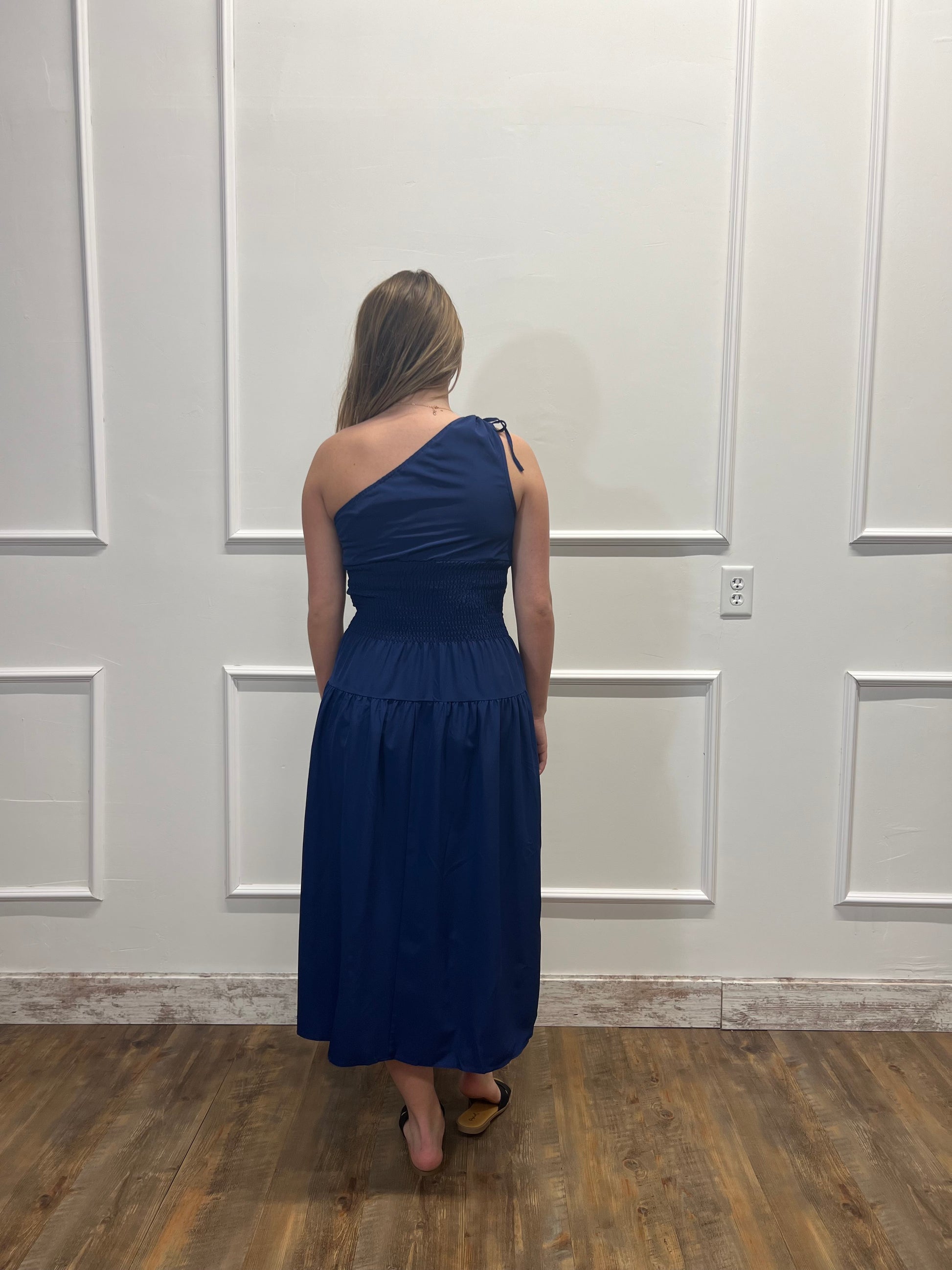 Woman wearing a blue one-shoulder dress standing against a white paneled wall.