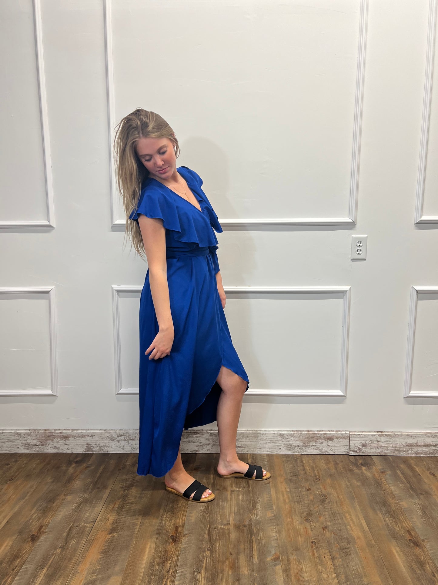 Woman in a blue dress standing against a white wall with wooden floor.