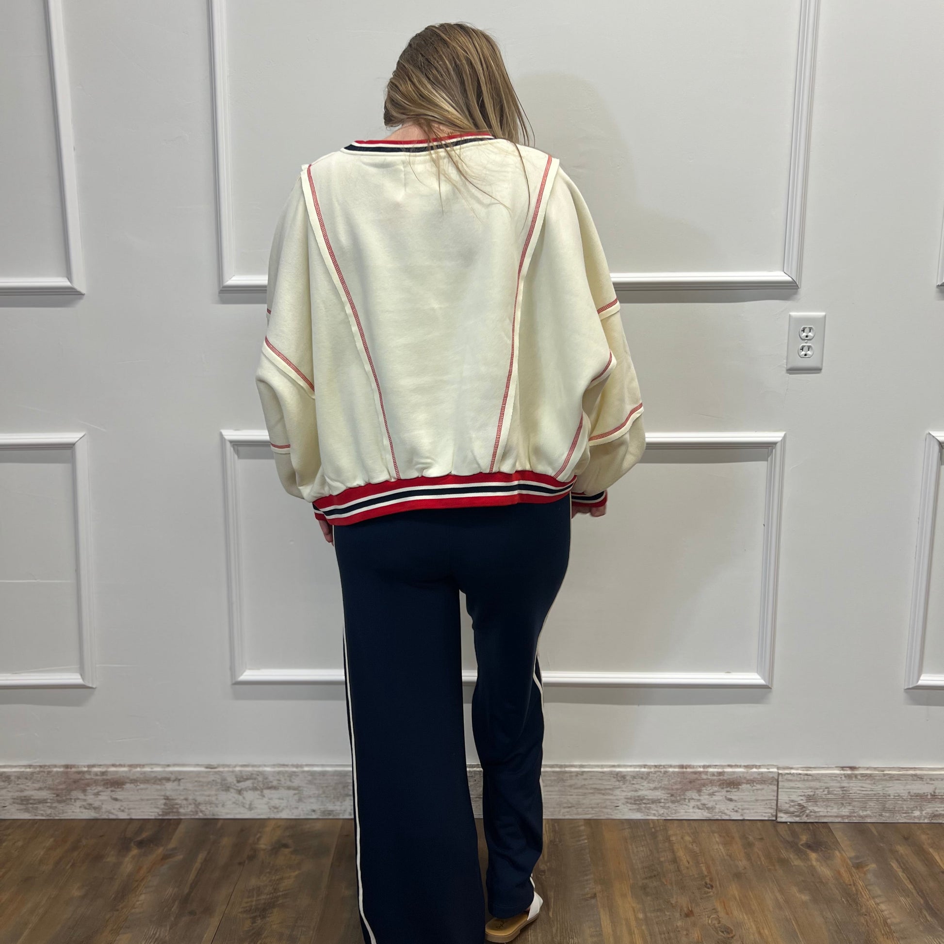 Person wearing a cream sweater with red trim and dark pants in a room with white paneled walls.