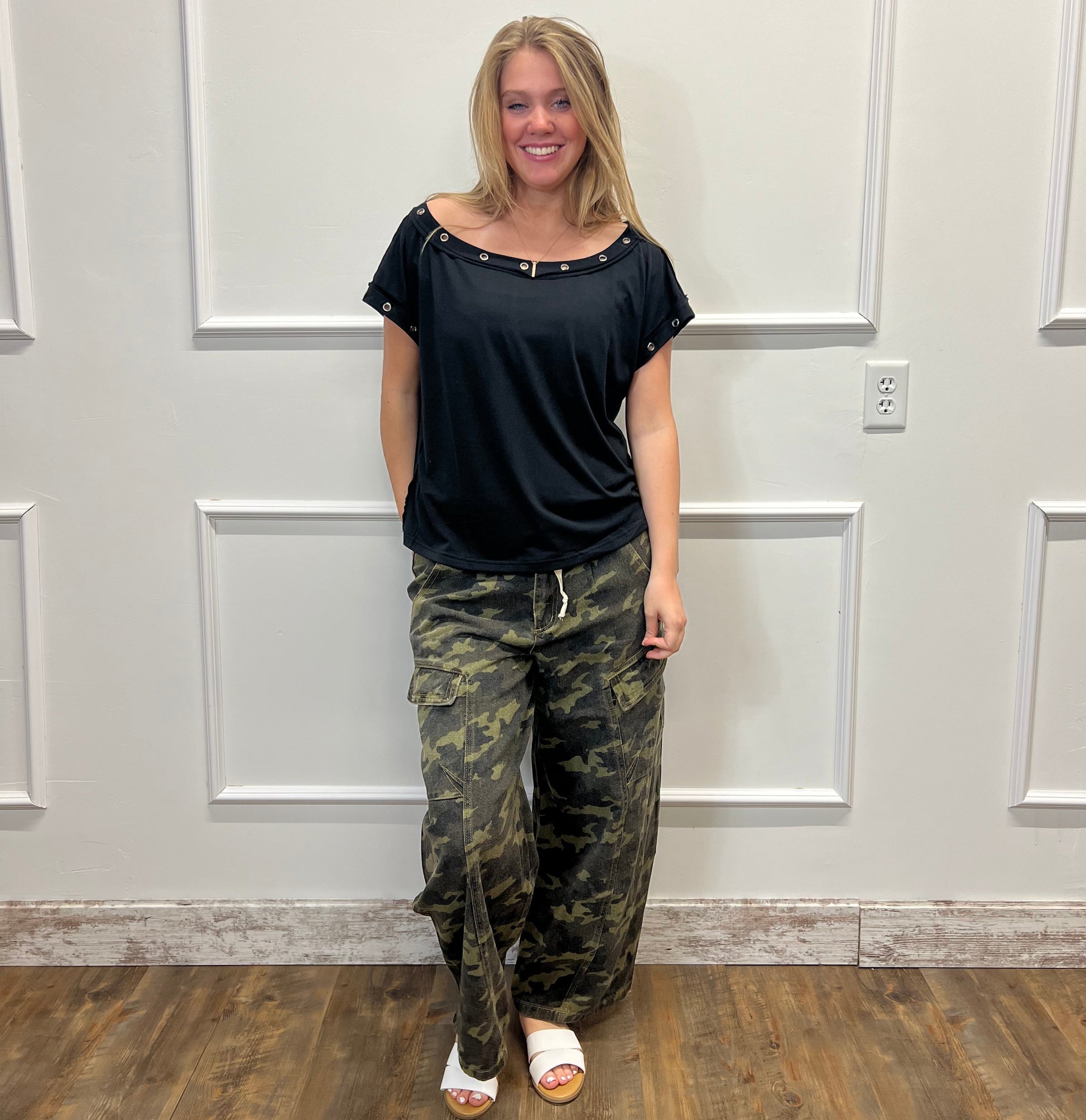 Person wearing a black top and camouflage pants standing in front of a white door.