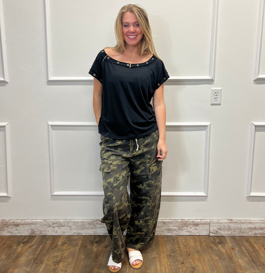 Person wearing a black top and camouflage pants standing in front of a white door.