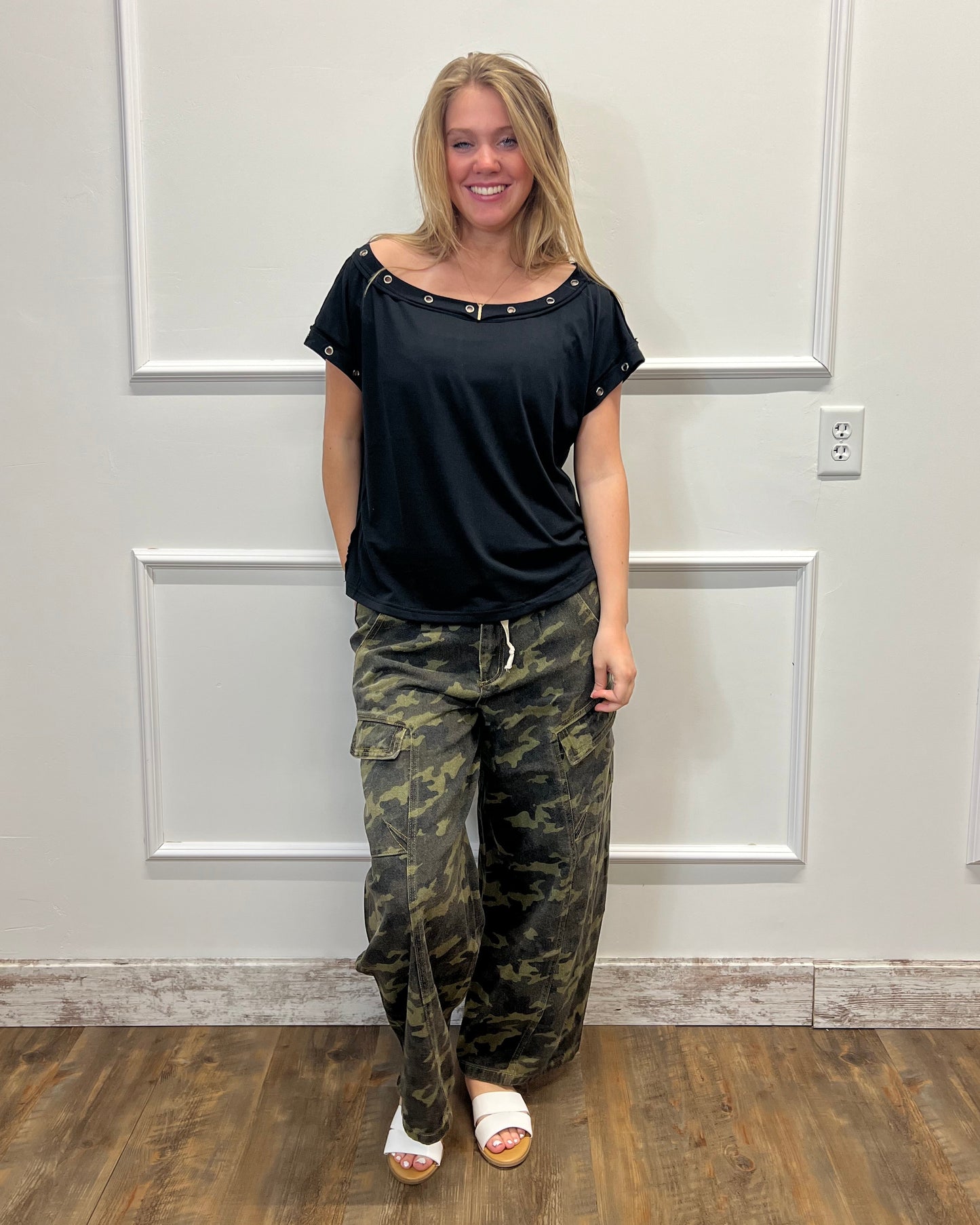 Person wearing a black top and camouflage pants standing in front of a white door.