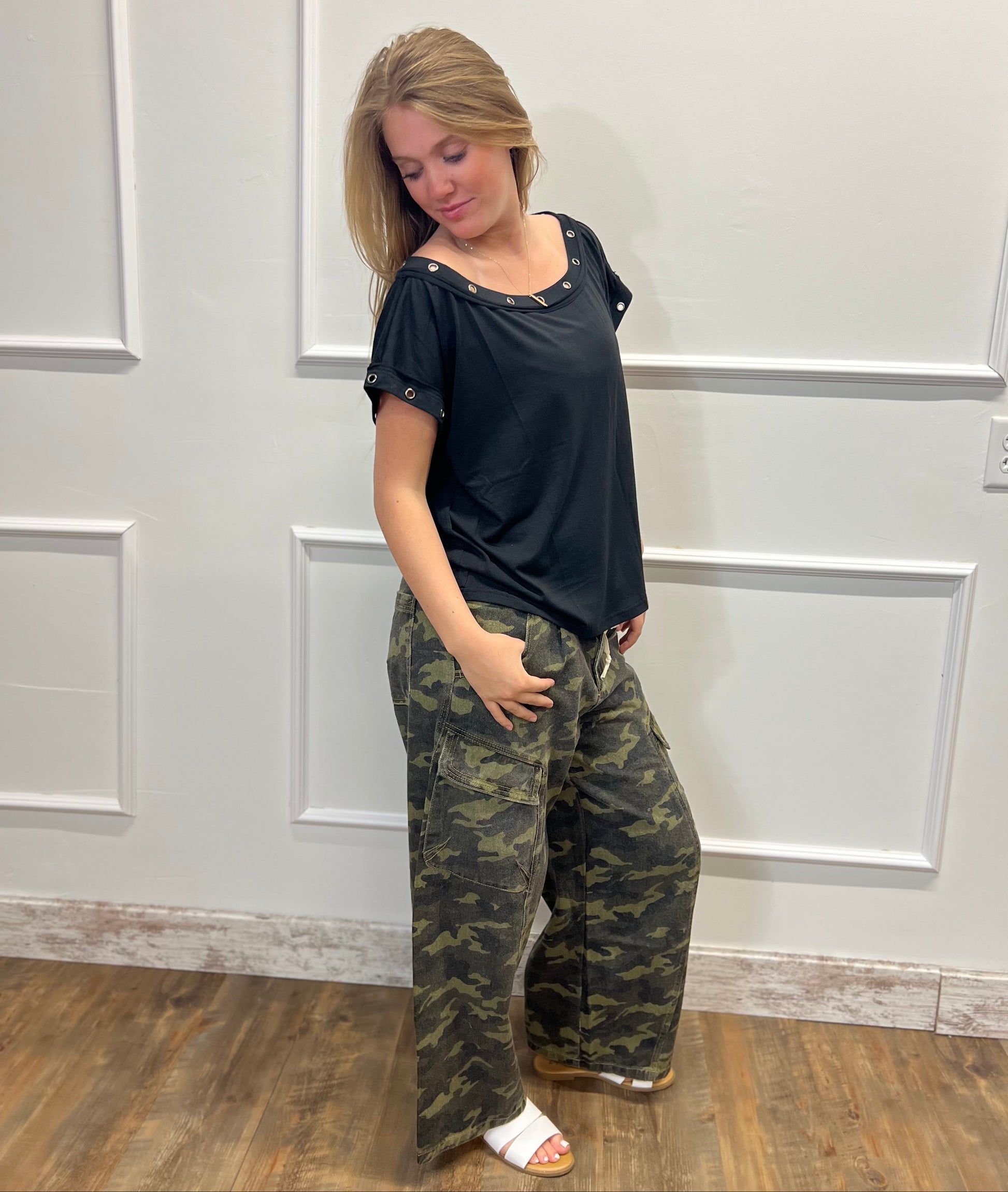 Woman wearing a black top and camouflage pants standing against a white paneled wall.