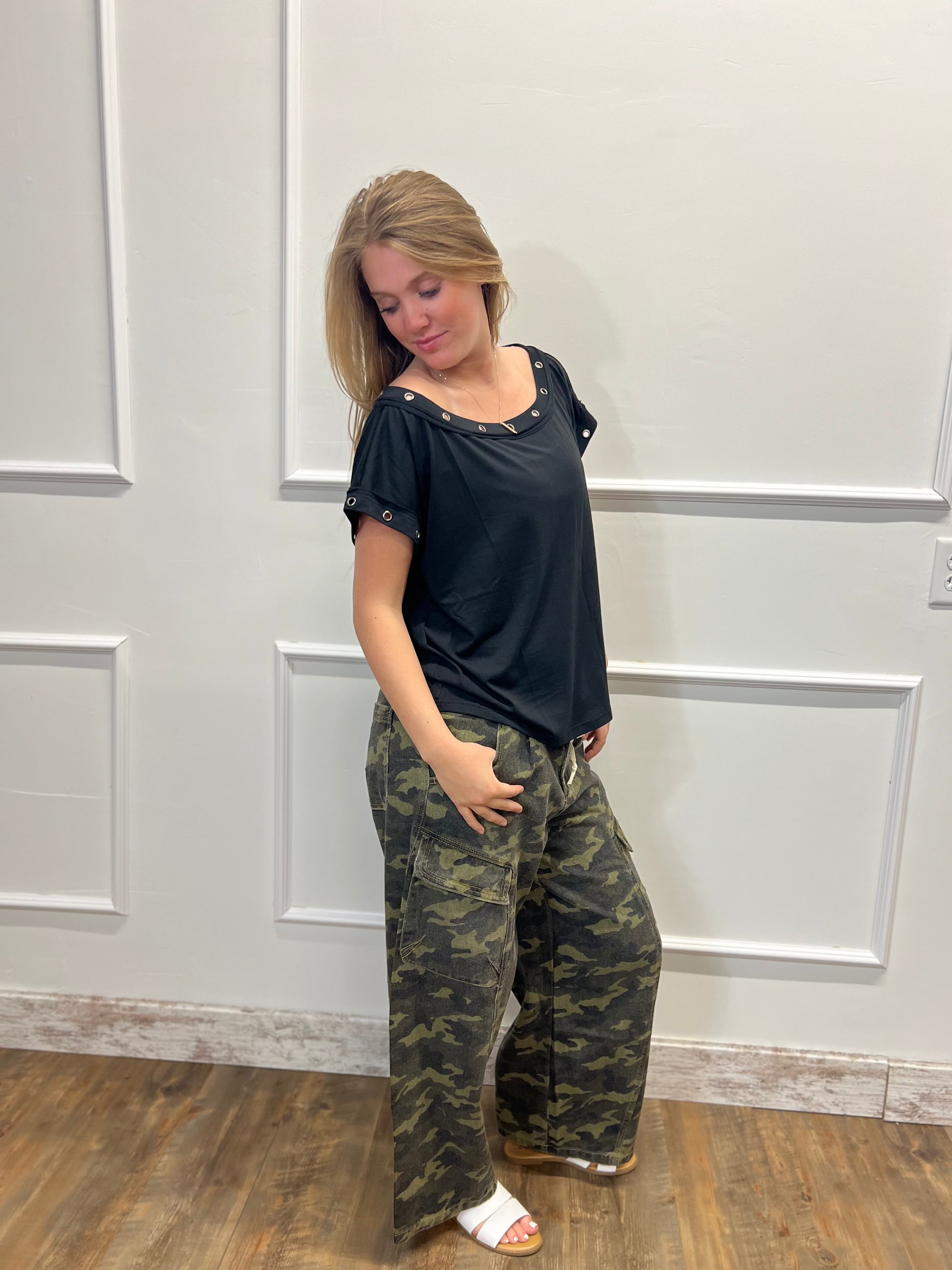 Woman wearing a black top and camouflage pants standing against a white paneled wall.