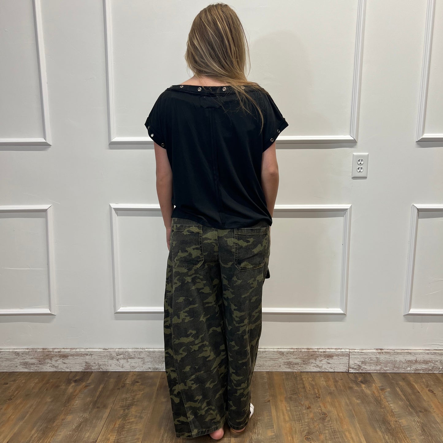 Person wearing a black shirt and camouflage pants standing in front of a white door.