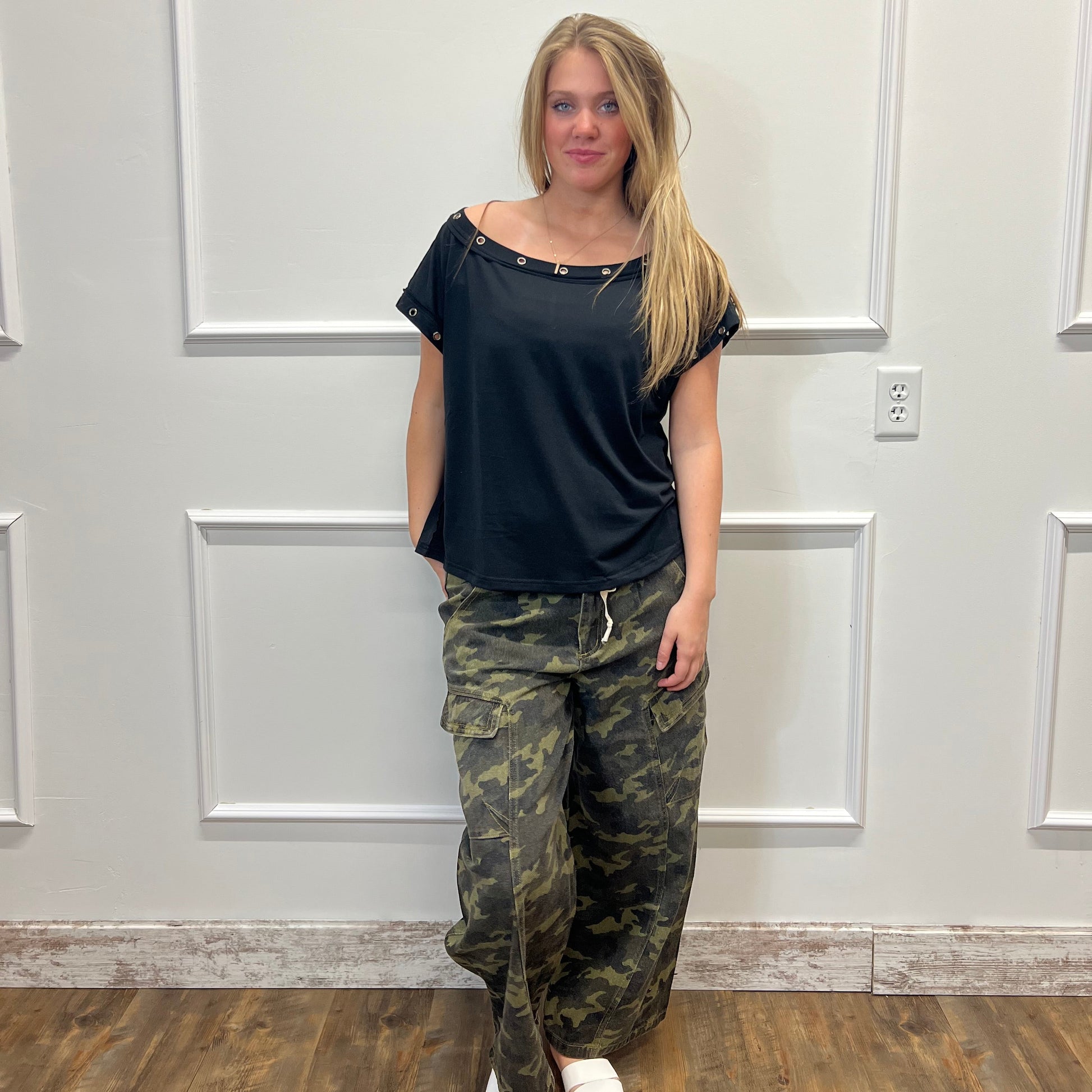 Person wearing a black top and camouflage pants standing in front of a white door.
