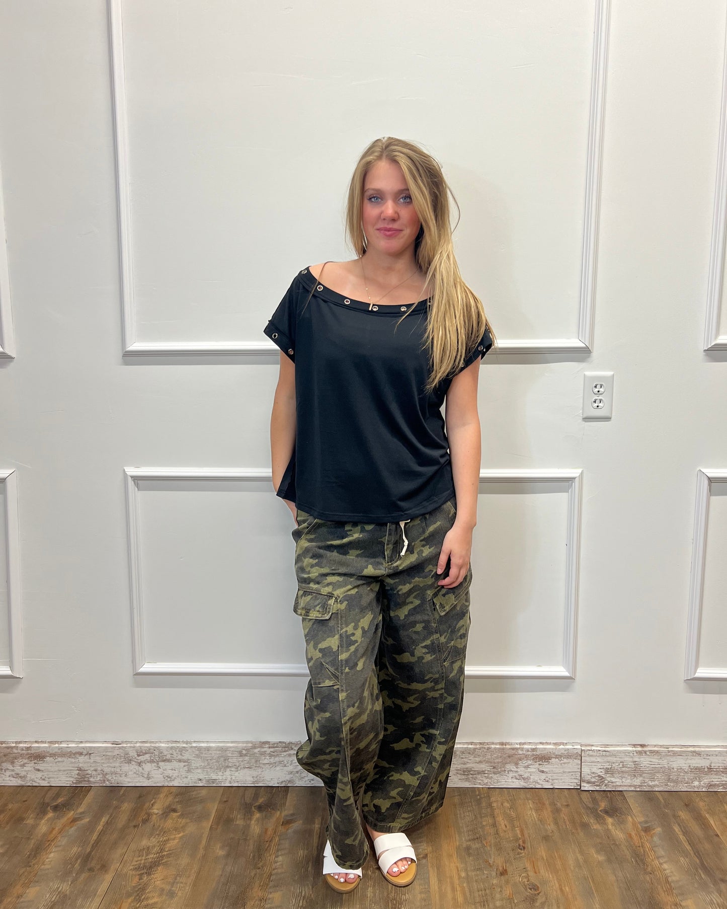 Person wearing a black top and camouflage pants standing in front of a white door.