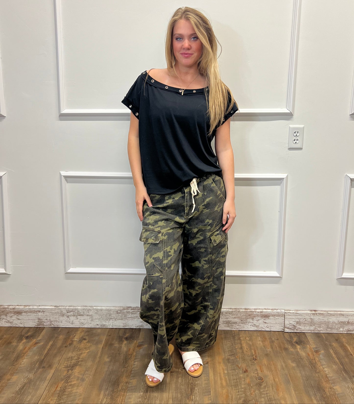 Person wearing a black top and camouflage pants standing in front of a white door.