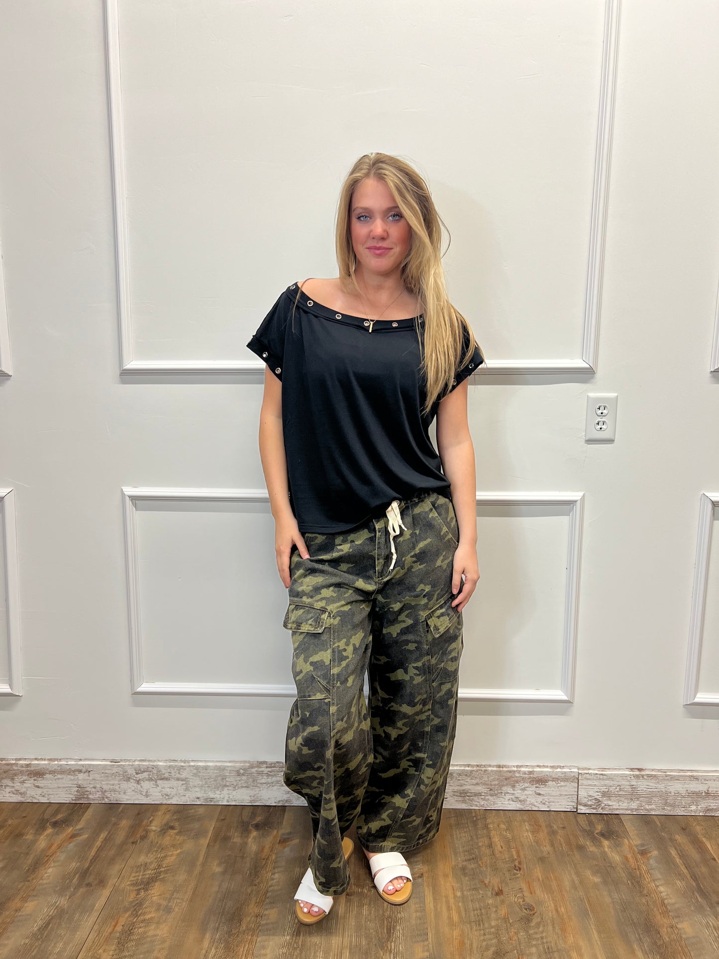 Person wearing a black top and camouflage pants standing in front of a white door.