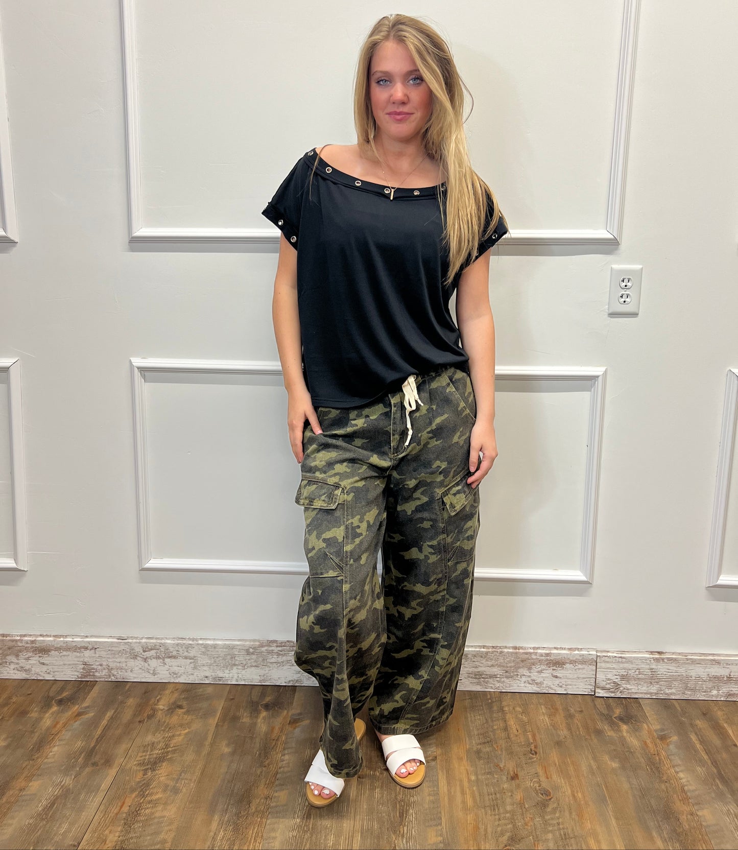 Person wearing a black top and camouflage pants standing in front of a white door.