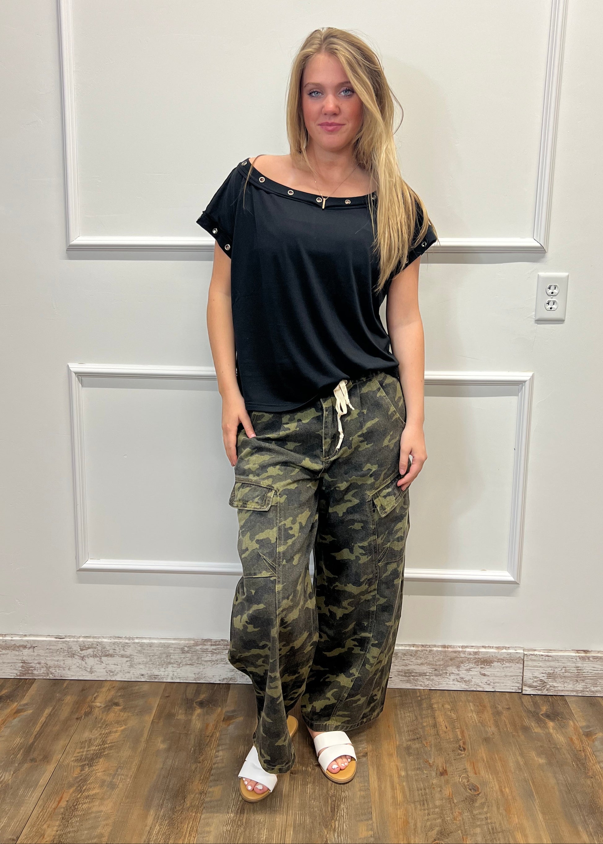 Person wearing a black top and camouflage pants standing in front of a white door.