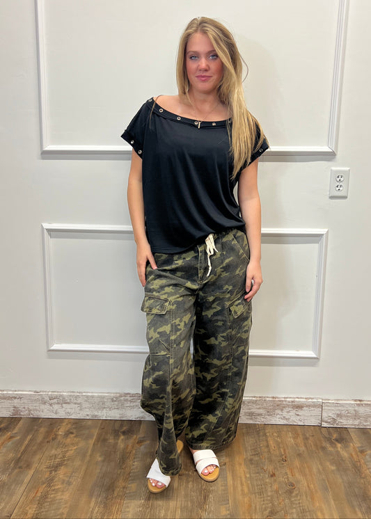 Person wearing a black top and camouflage pants standing in front of a white door.