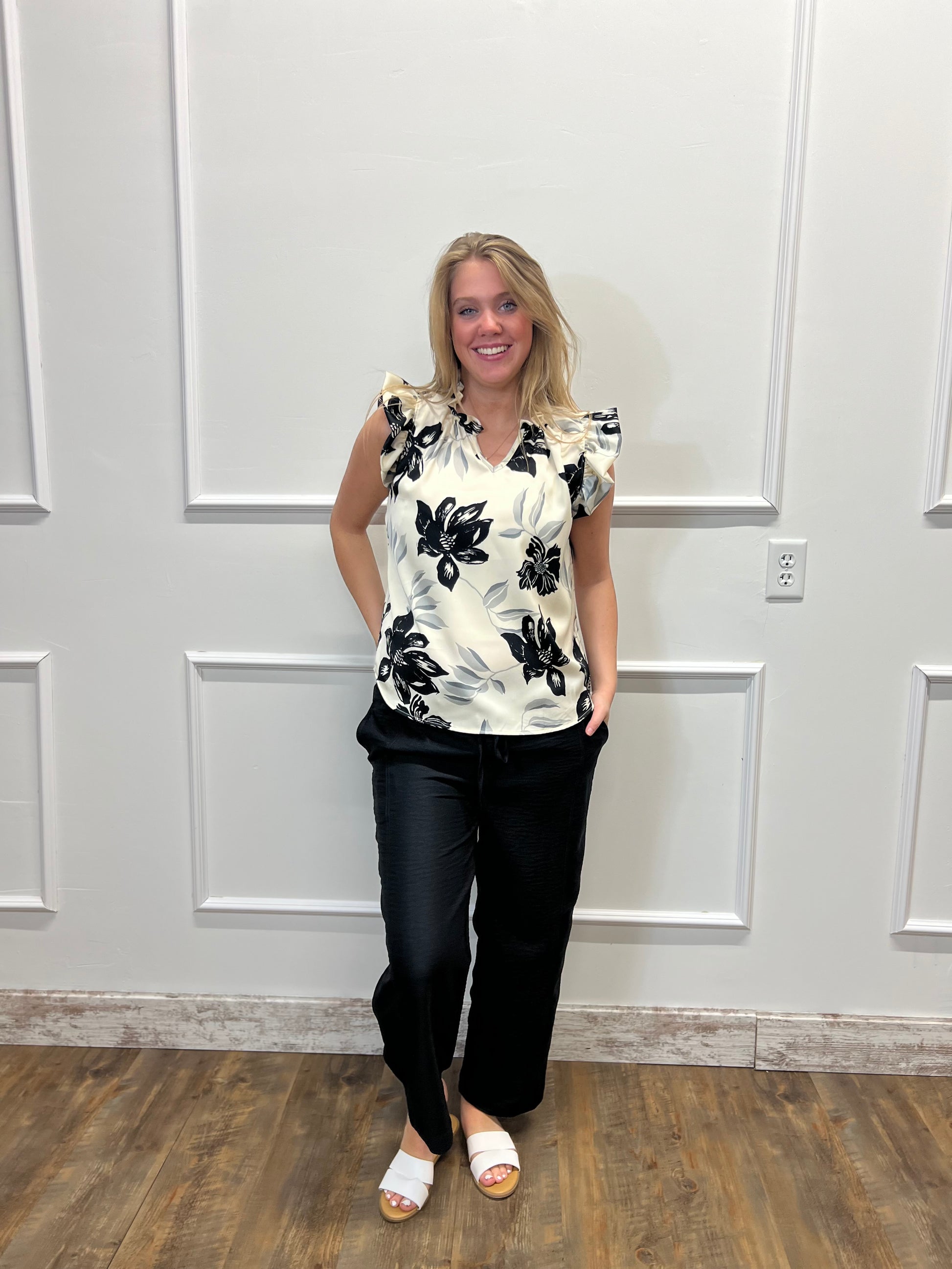 Woman wearing a black and white floral top and black pants standing against a white paneled wall.