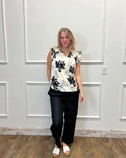 Woman wearing a floral top and black pants against a white paneled wall.