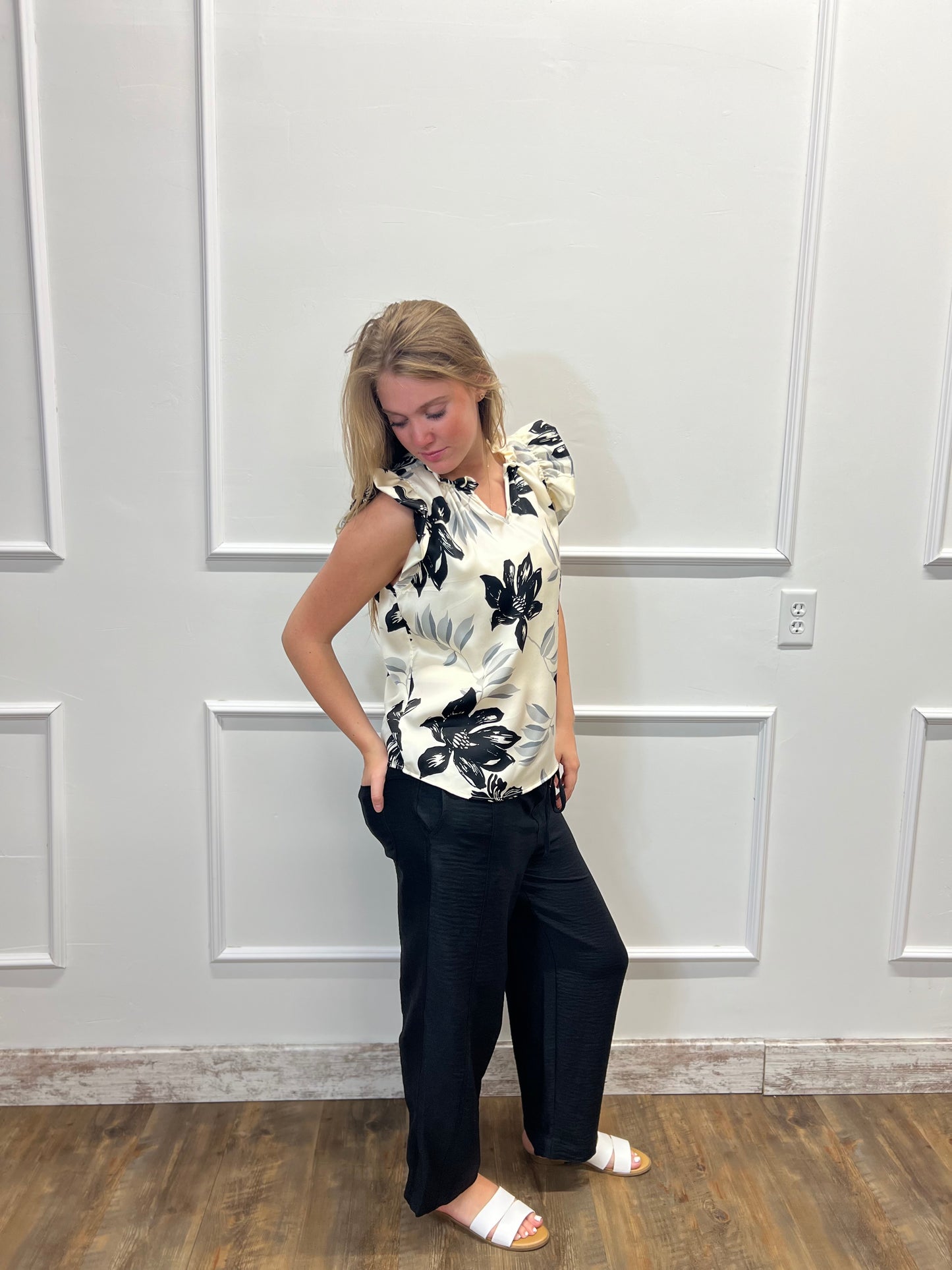 Woman wearing a floral top and black pants against a white paneled wall.