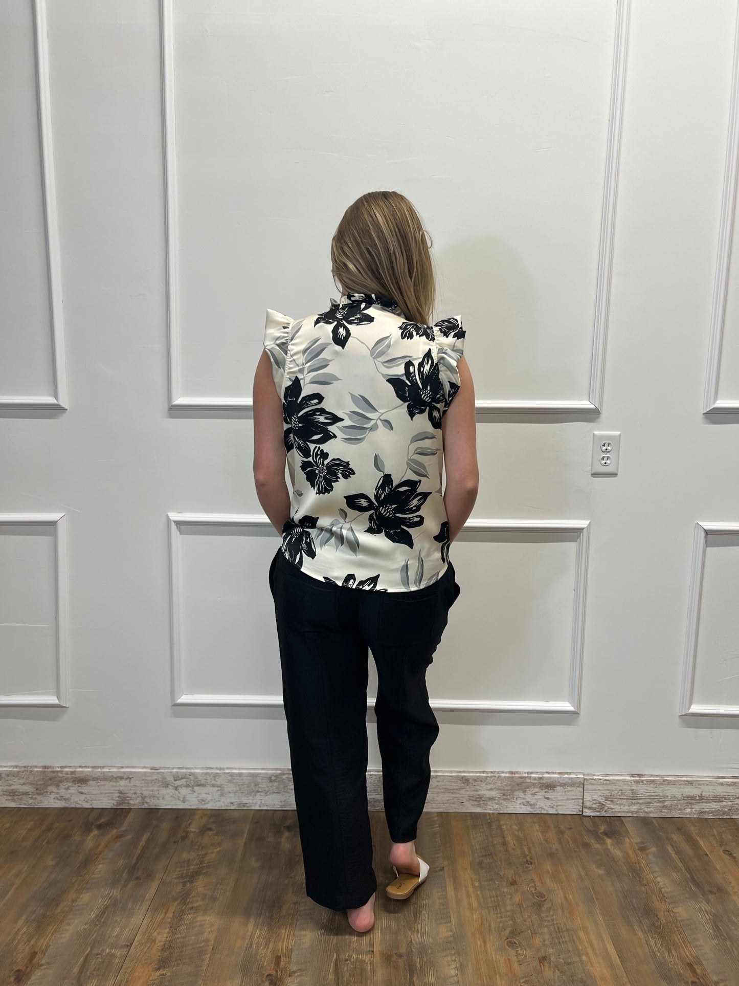 Person wearing a sleeveless floral top and black pants standing against a white paneled wall.