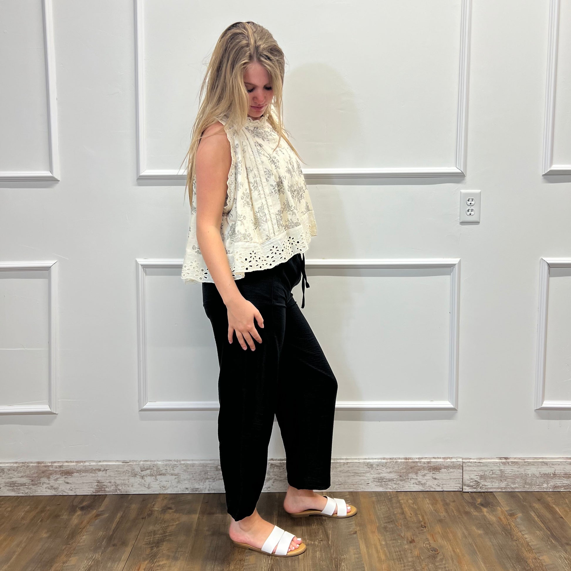 Woman wearing a white sleeveless top and black pants standing in a room with white paneled walls.