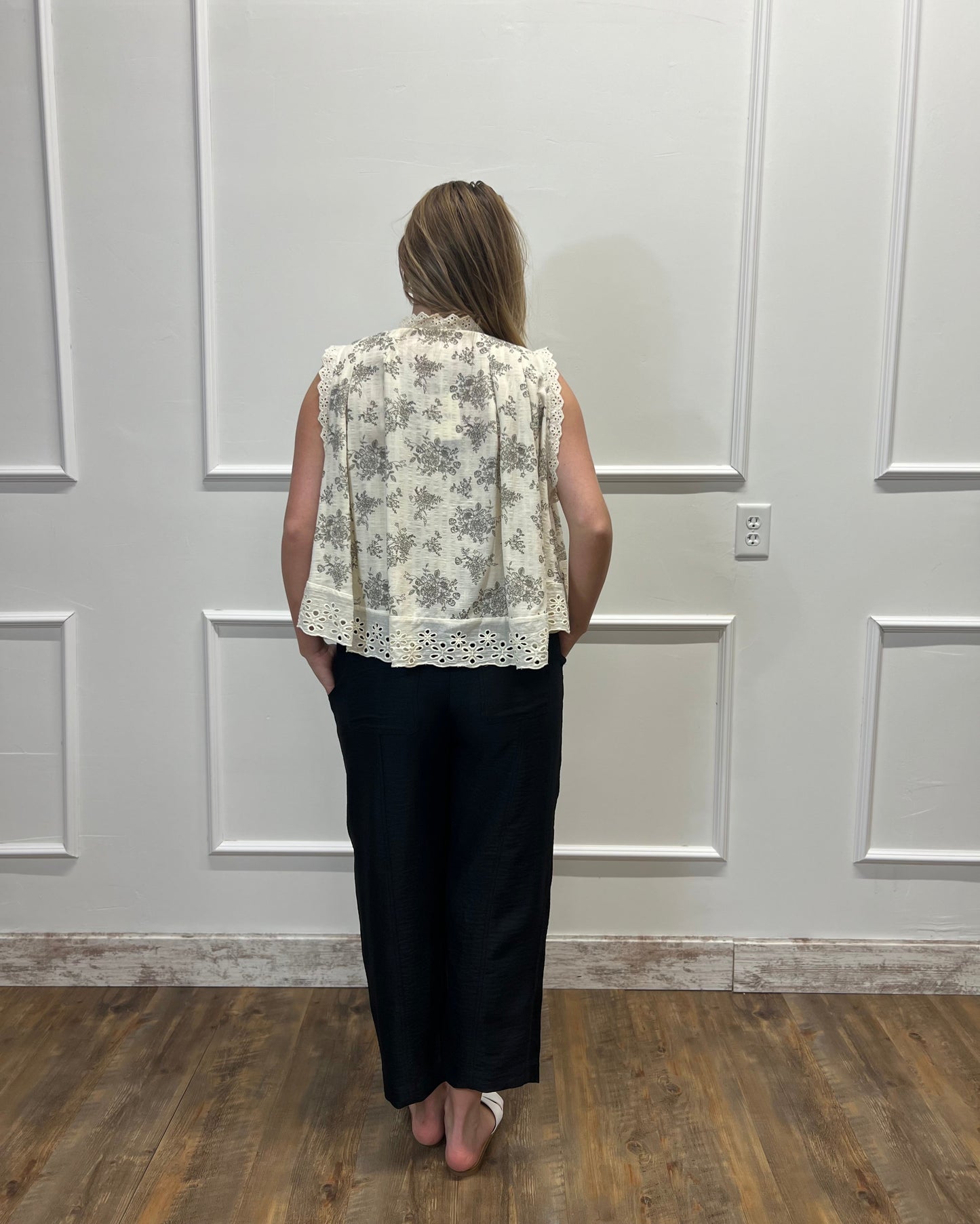 Person wearing a floral blouse and black pants standing against a white paneled wall.