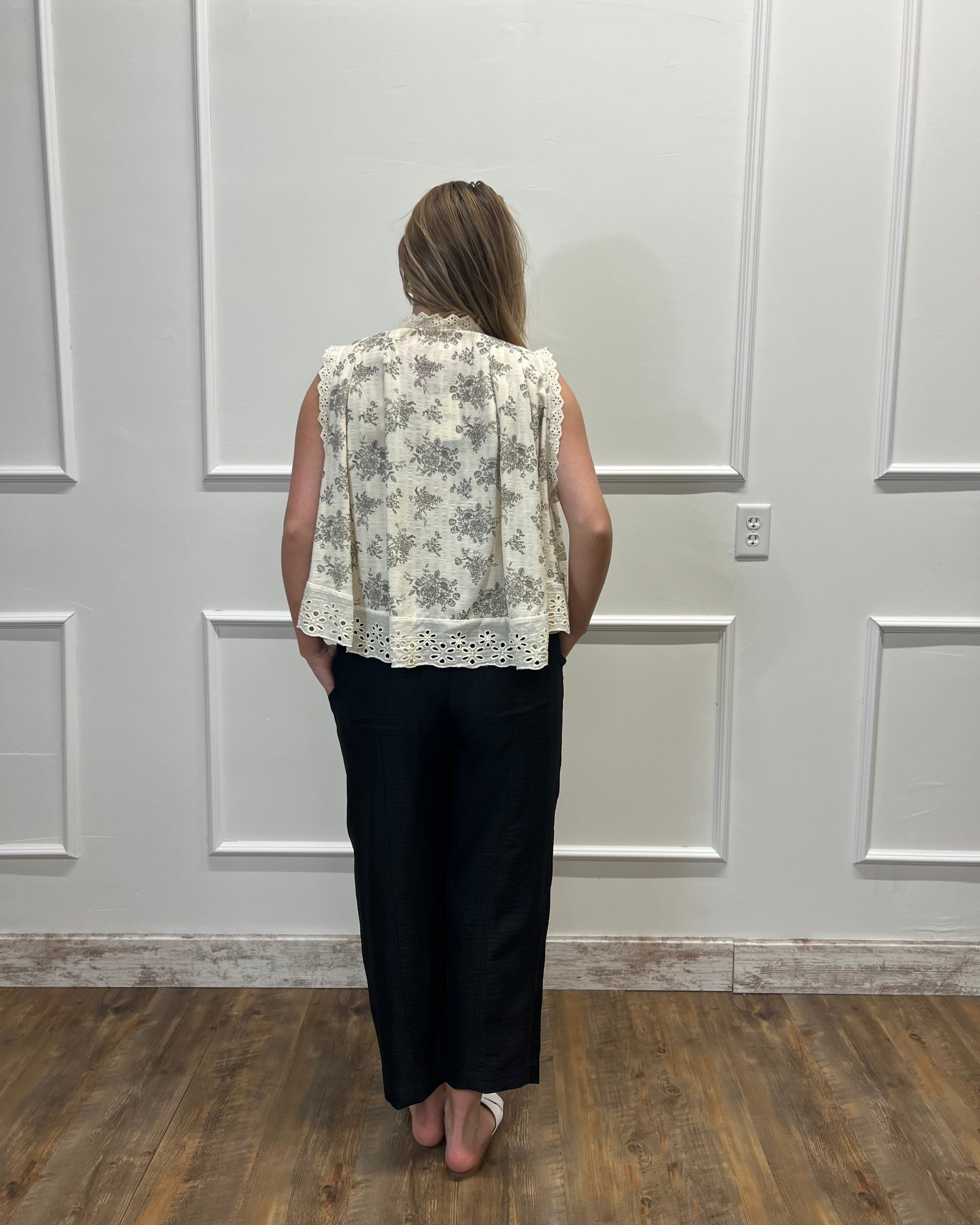 Person wearing a floral blouse and black pants standing against a white paneled wall.
