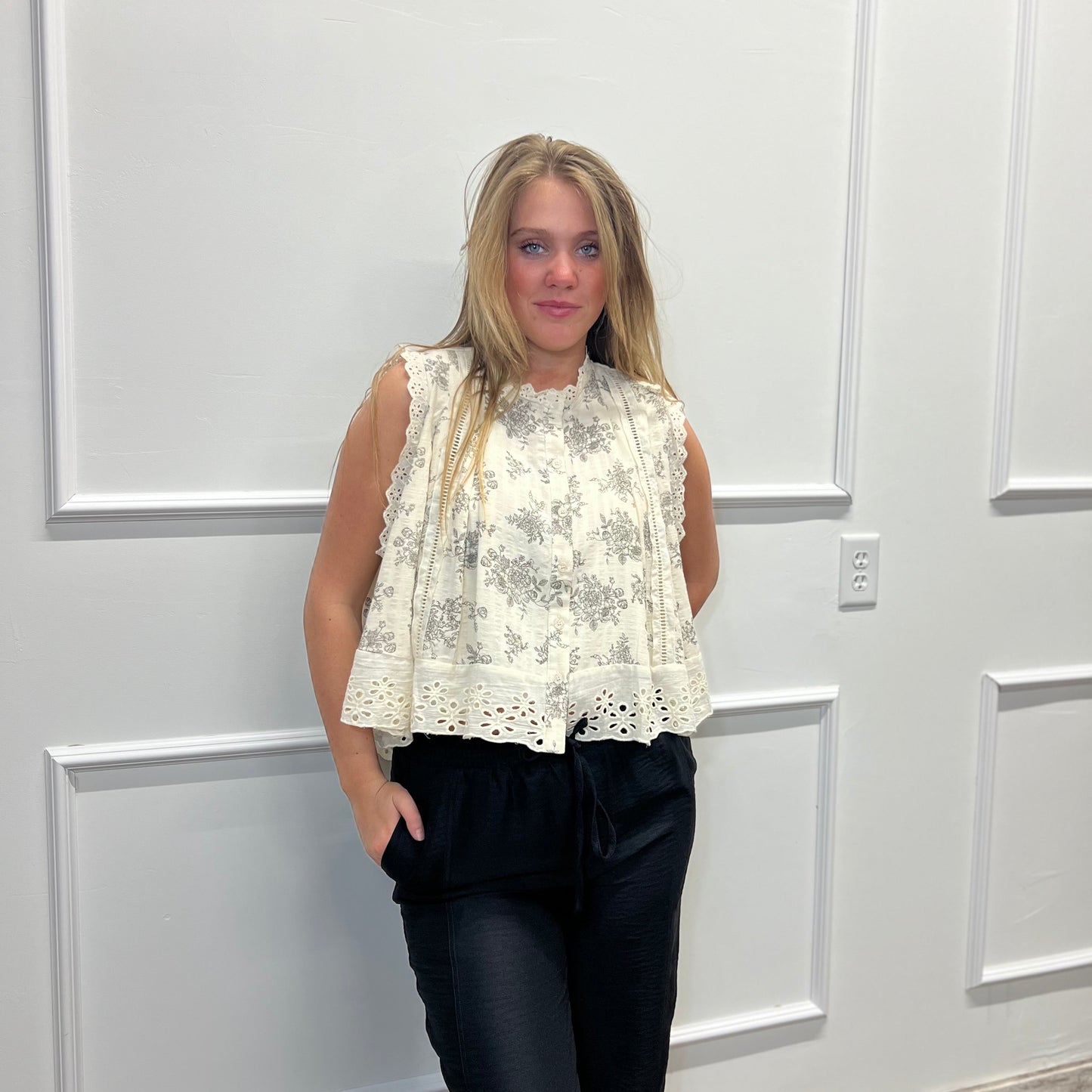 Woman wearing a white lace top and black pants standing against a white paneled wall.
