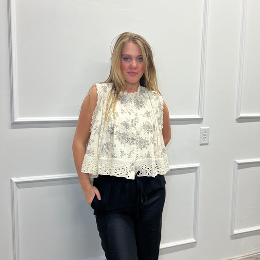 Woman wearing a white lace top and black pants standing against a white paneled wall.
