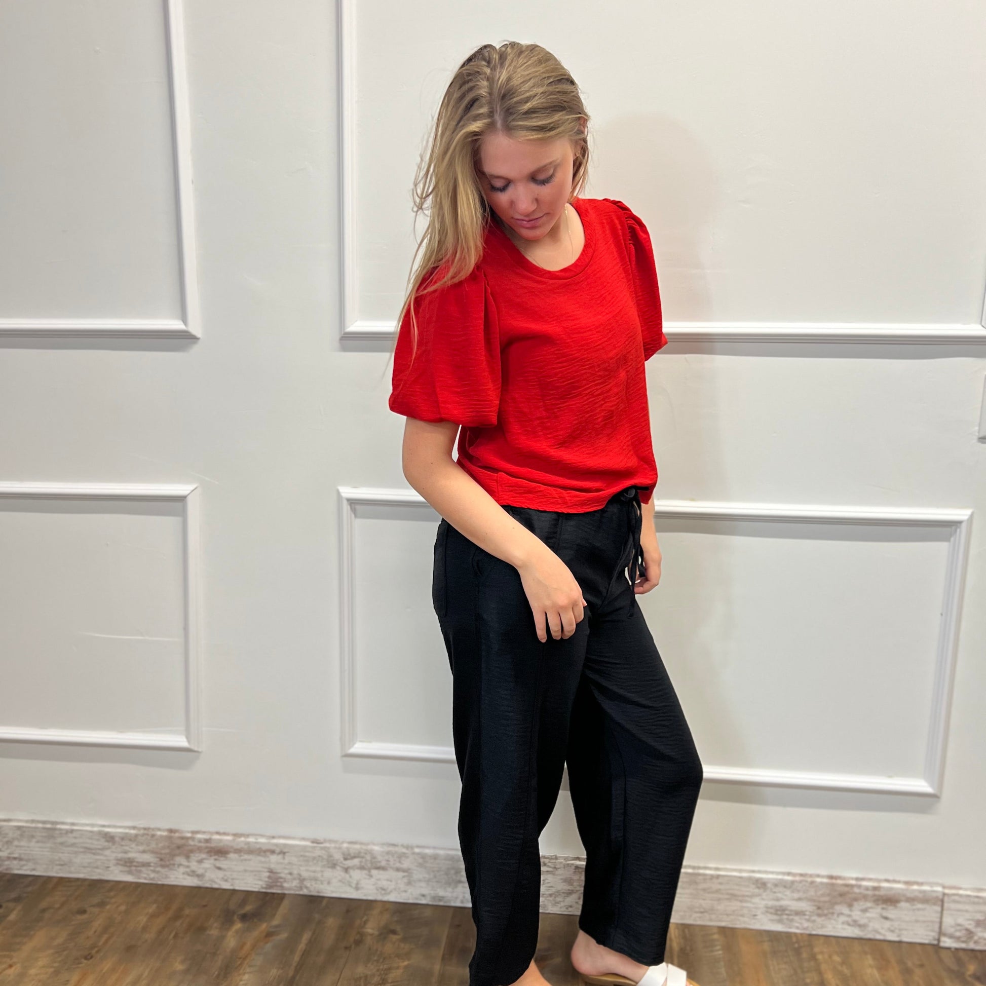 Woman in a red top and black pants standing against a white paneled wall.