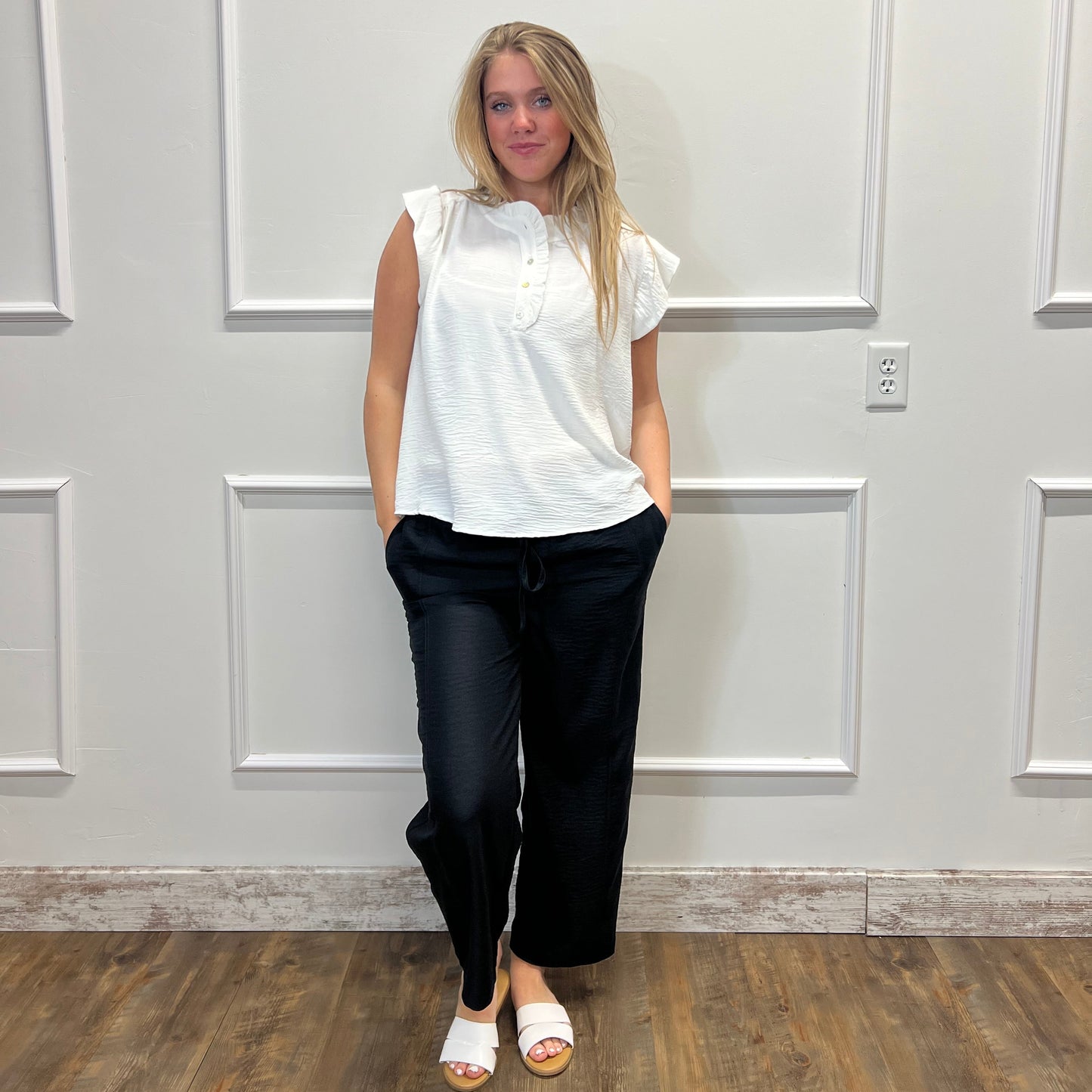 Woman wearing a white blouse and black pants standing against a white paneled wall.